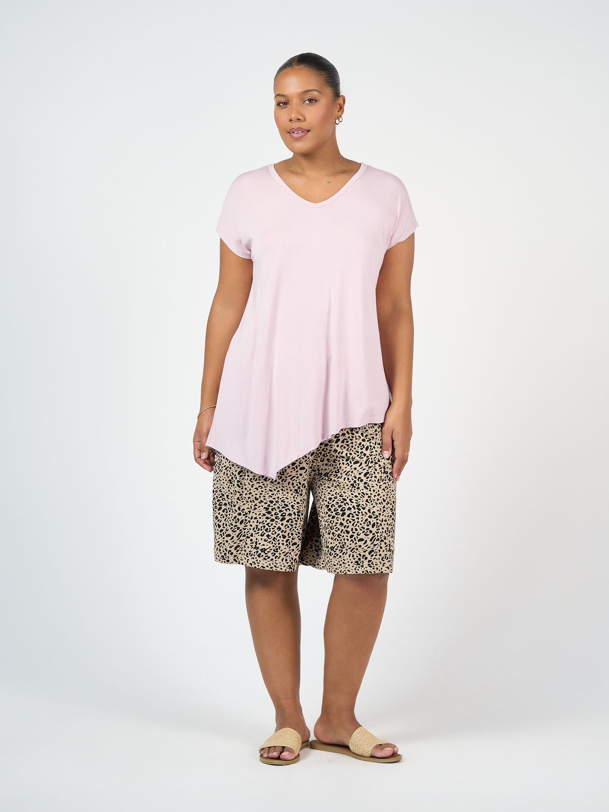 A woman stands against a plain light background wearing the Holiday Asymmetrical Bamboo V-Neck T-Shirt in Lilac, paired with leopard print knee-length shorts, gold hoop earrings, and beige sandals.
