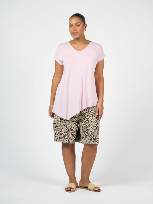 A woman stands against a plain light background wearing the Holiday Asymmetrical Bamboo V-Neck T-Shirt in Lilac, paired with leopard print knee-length shorts, gold hoop earrings, and beige sandals.