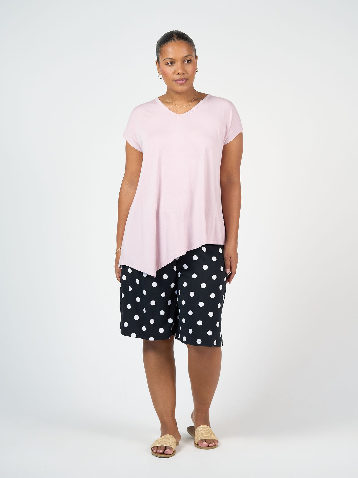 A woman stands against a plain background wearing the Holiday Asymmetrical Bamboo V-Neck T-Shirt in lilac, black knee-length shorts with white polka dots, tan slide sandals, and hoop earrings.