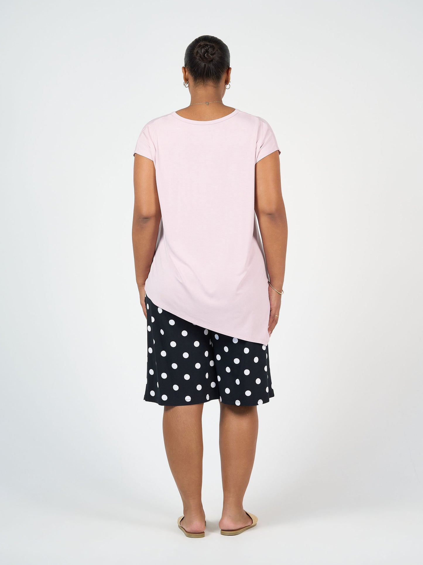 A woman faces away from the camera, wearing the Holiday Asymmetrical Bamboo V-Neck T-Shirt in Lilac, black polka dot shorts, and sandals against a plain white background.