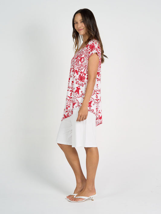 A woman stands sideways, smiling, wearing the Holiday Asymmetrical Bamboo V-Neck T-Shirt in Lipstick Palm, paired with white knee-length shorts and white heeled sandals against a plain white background.