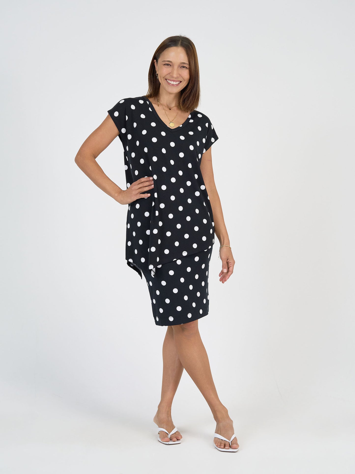 A woman smiles in the Midi Bamboo Whitney Tube Skirt - Polka Dot, a slimming black skirt with white polka dots and an asymmetrical hem. She pairs it with white sandals and poses against a plain white background.