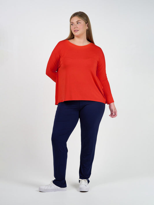 A woman with long, straight hair stands against a plain white background, wearing the Warwick Bamboo Fold Over Waist Pant in Navy and a bright red long-sleeve top. She looks slightly to the side with a relaxed expression.