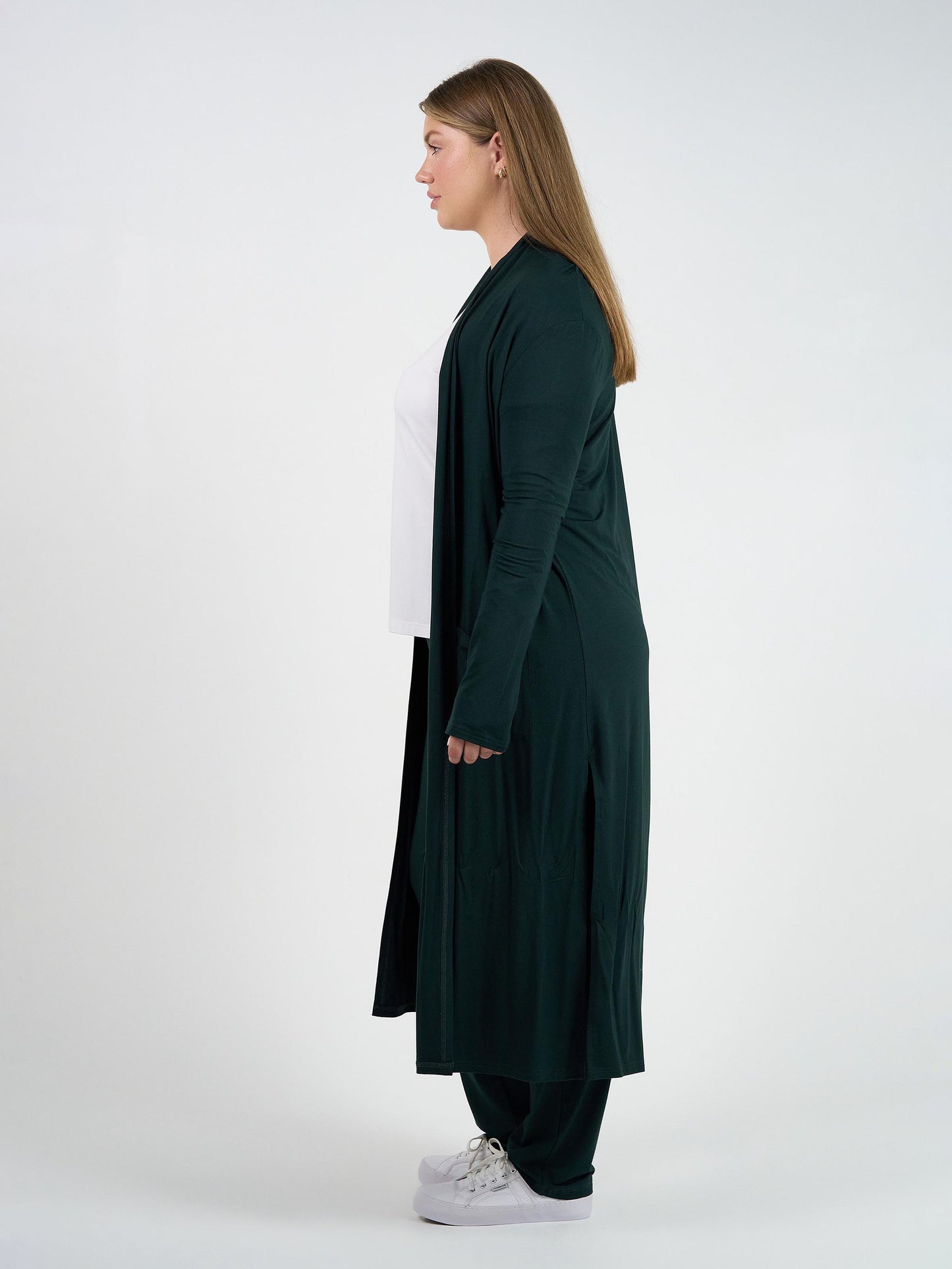 A woman stands in profile against a plain white background, wearing the Bamboo Longline Pocket Cardigan - Dark Green over a white top, matching pants, and white sneakers—a sustainable essential ideal for travel.