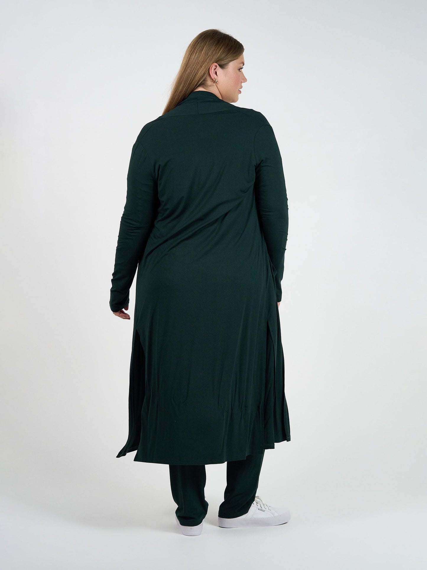 A woman stands facing away, wearing the Bamboo Longline Pocket Cardigan in dark green—an ideal sustainable wardrobe essential—over matching pants and white sneakers. The relaxed-fit cardigan with side slits is set against a plain white background.