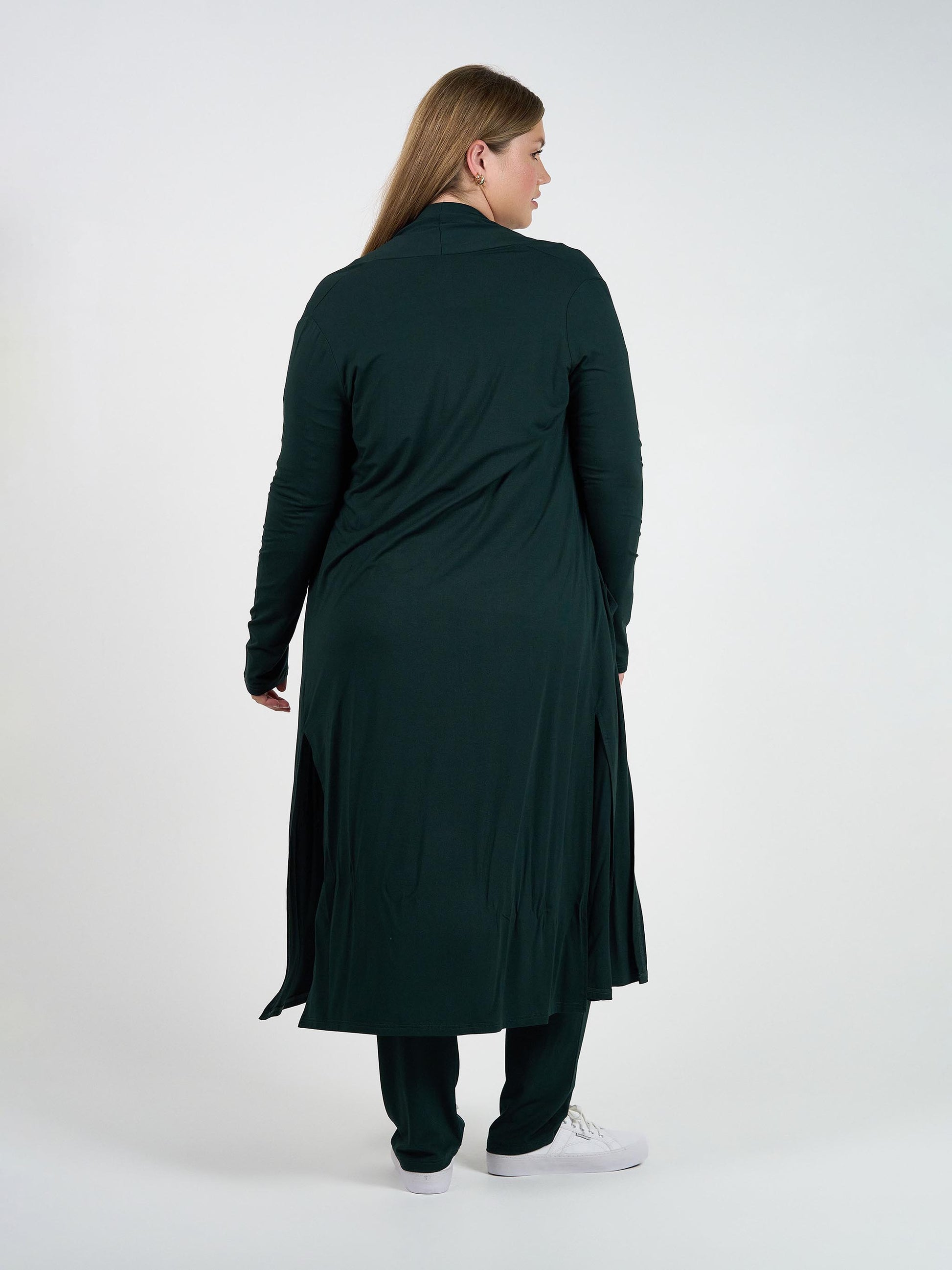 A woman stands facing away, wearing the Bamboo Longline Pocket Cardigan in dark green—an ideal sustainable wardrobe essential—over matching pants and white sneakers. The relaxed-fit cardigan with side slits is set against a plain white background.