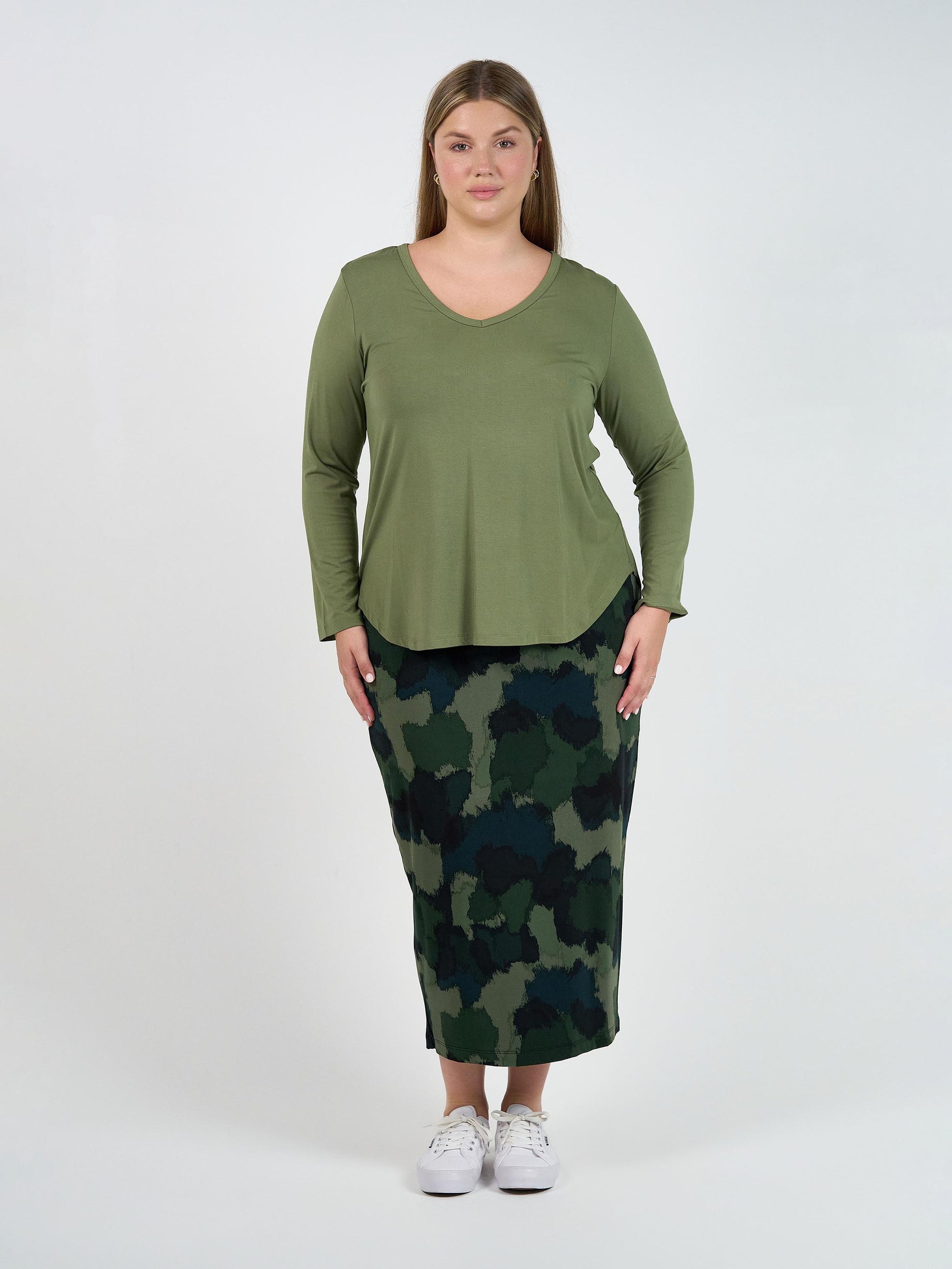 A woman faces forward against a plain white background, wearing the Sia V Neck Bamboo Longsleeve Top in olive green, paired with a dark green camo skirt and white sneakers.