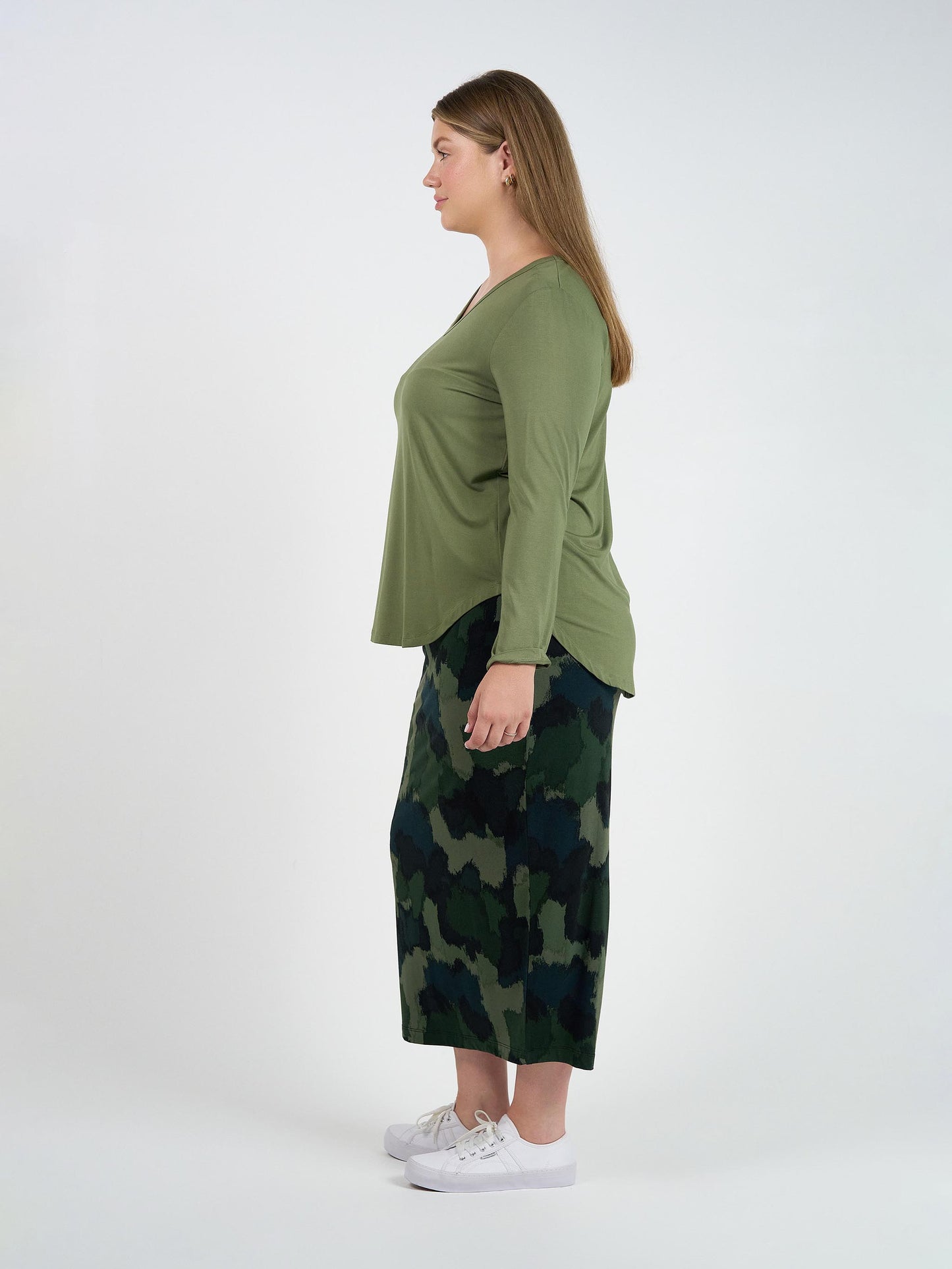 A woman stands in profile against a white background, wearing the Sia V Neck Bamboo Longsleeve Top in olive green, paired with a green camo-patterned skirt and white sneakers.