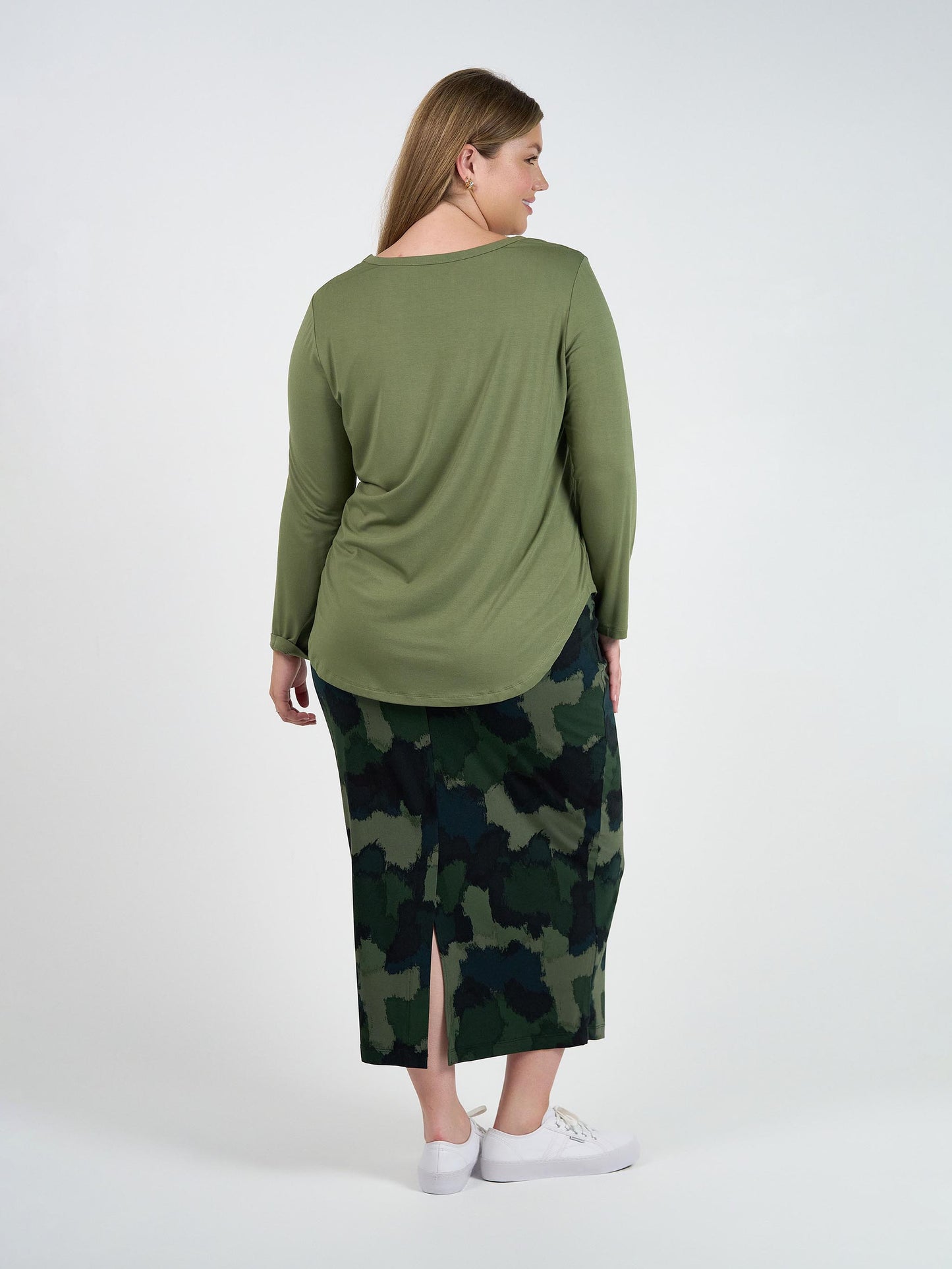 A woman with long blonde hair, seen from behind, wears the Sia V Neck Bamboo Longsleeve Top in olive green, a camouflage skirt with a back slit, and white sneakers against a plain white background.