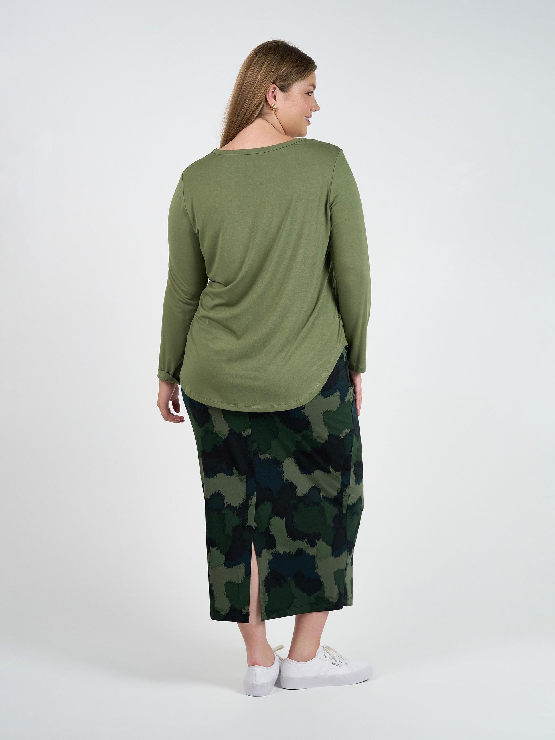 A woman with long blonde hair, seen from behind, wears the Sia V Neck Bamboo Longsleeve Top in olive green, a camouflage skirt with a back slit, and white sneakers against a plain white background.