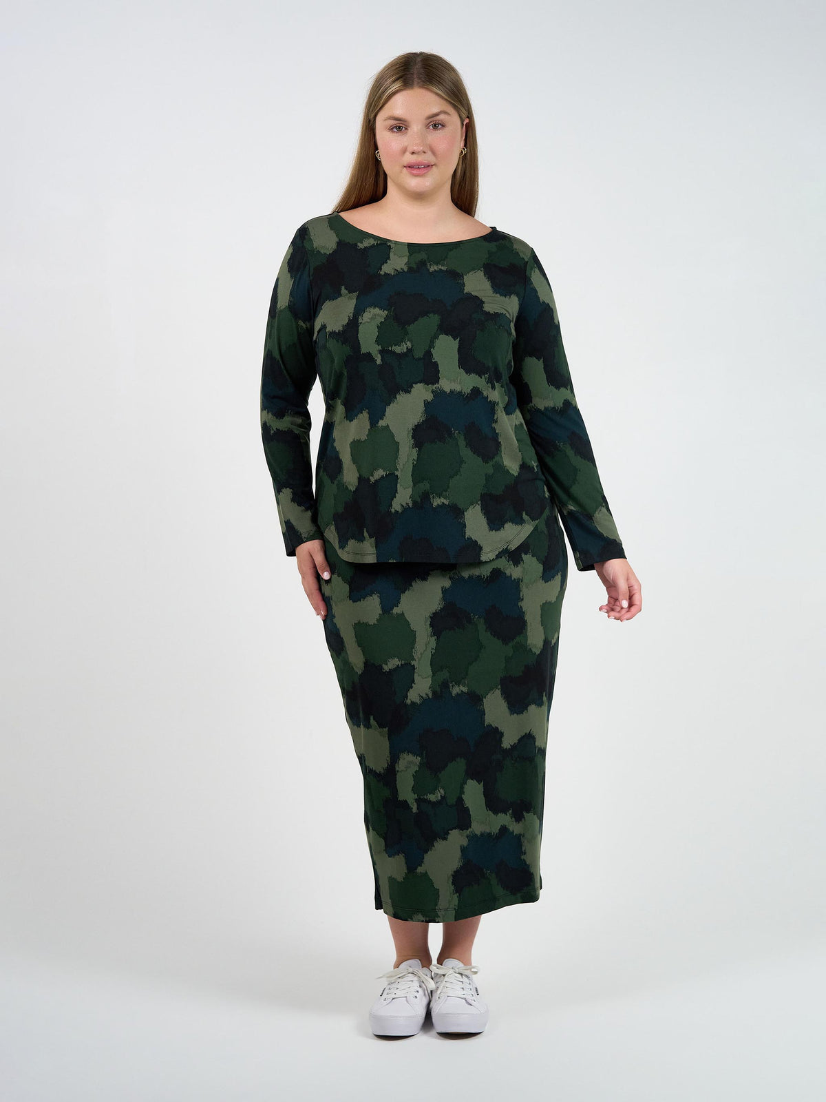 A woman with long, light brown hair stands against a white background, wearing the Adele Long Sleeve Bamboo Top in camouflage print with a matching skirt and white sneakers, looking relaxed.