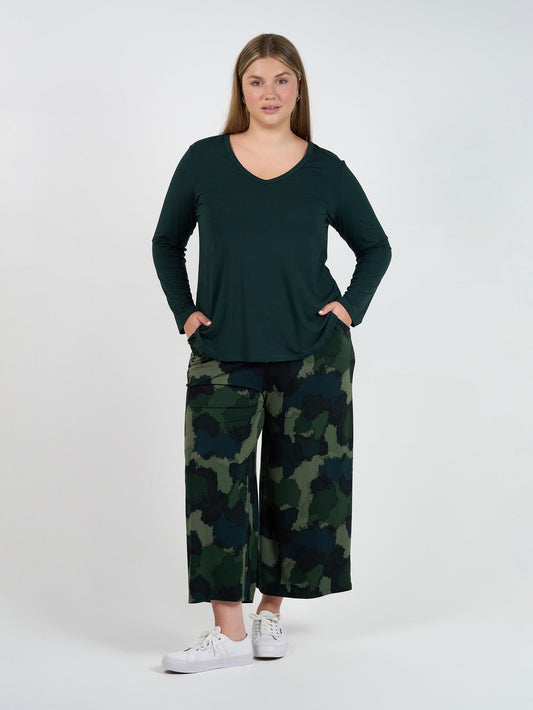 A woman stands against a white background wearing a long-sleeve dark green top, white sneakers, and Ciara 7/8 Length Bamboo Culottes in a camouflage print, with her hands in her pockets and a neutral expression.
