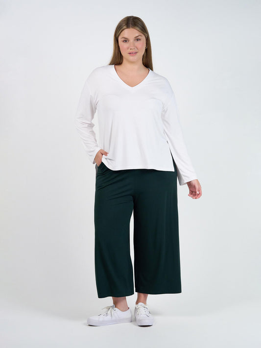 A woman stands against a plain white background wearing a long-sleeve white V-neck top, Ciara 7/8 Length Bamboo Culottes in dark green, and white sneakers, with one hand in her pocket.