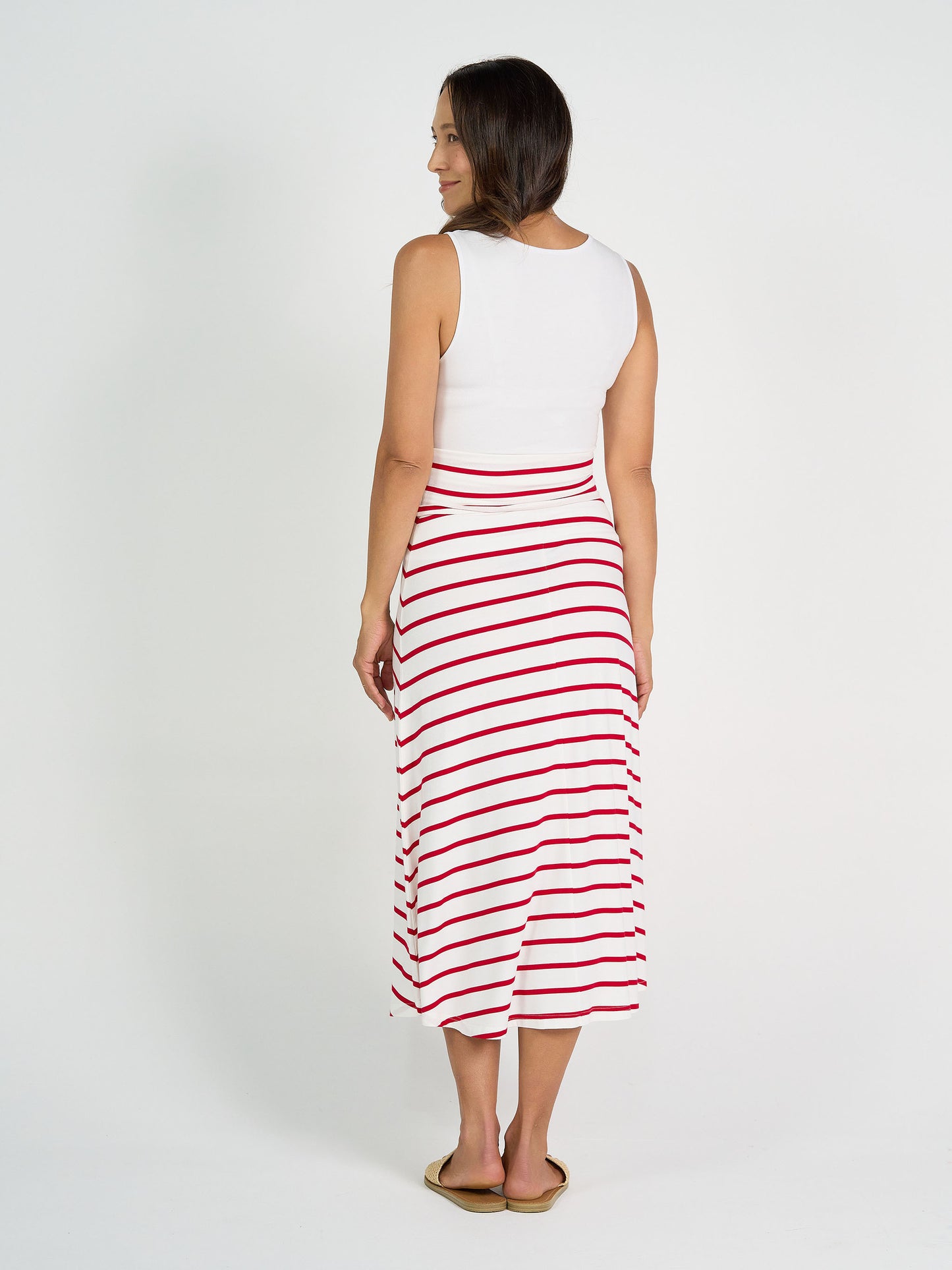 A woman faces away, wearing a sleeveless white top, tan sandals, and the Jackson Bamboo Maxi Skirt in Lipstick & White Stripe, against a plain white background.