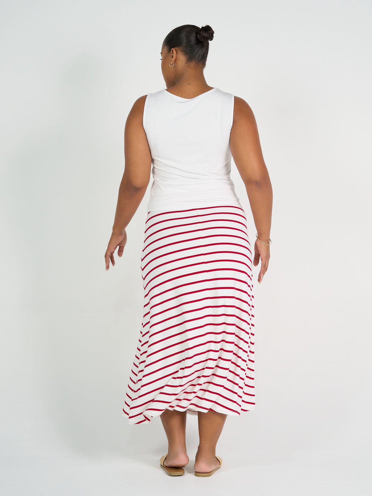A person with a bun, in a sleeveless white top and the Jackson Bamboo Maxi Skirt - Lipstick & White Stripe, stands barefoot facing away against a plain white background.