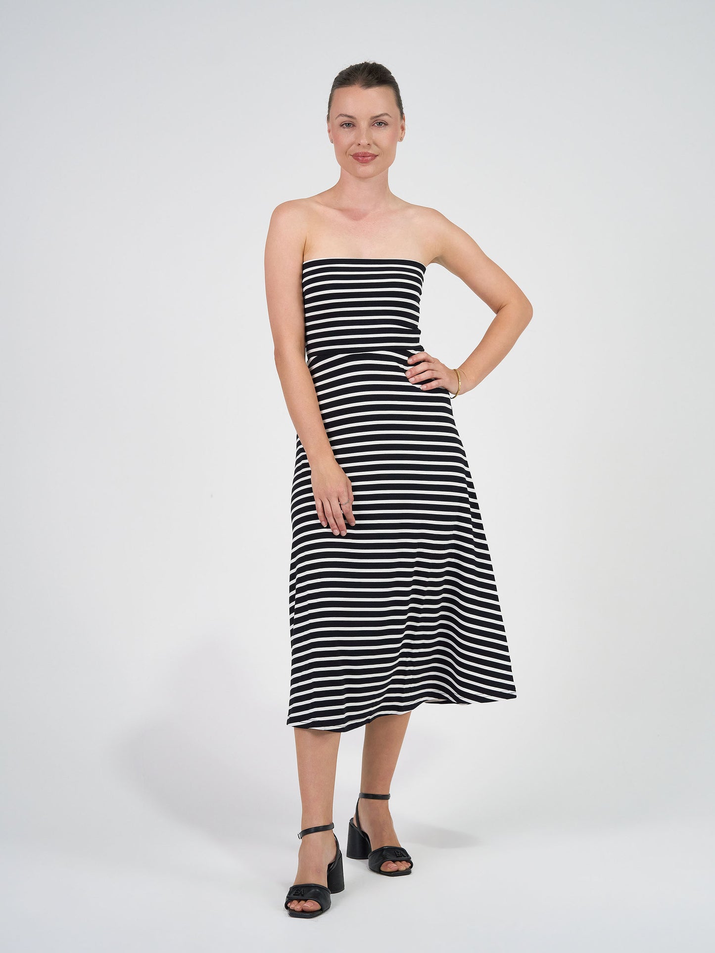 A woman stands against a white background, smiling with one hand on her hip. She wears the Jackson Bamboo Maxi Skirt in Black & White Stripe and black heeled sandals, with her hair tied back.