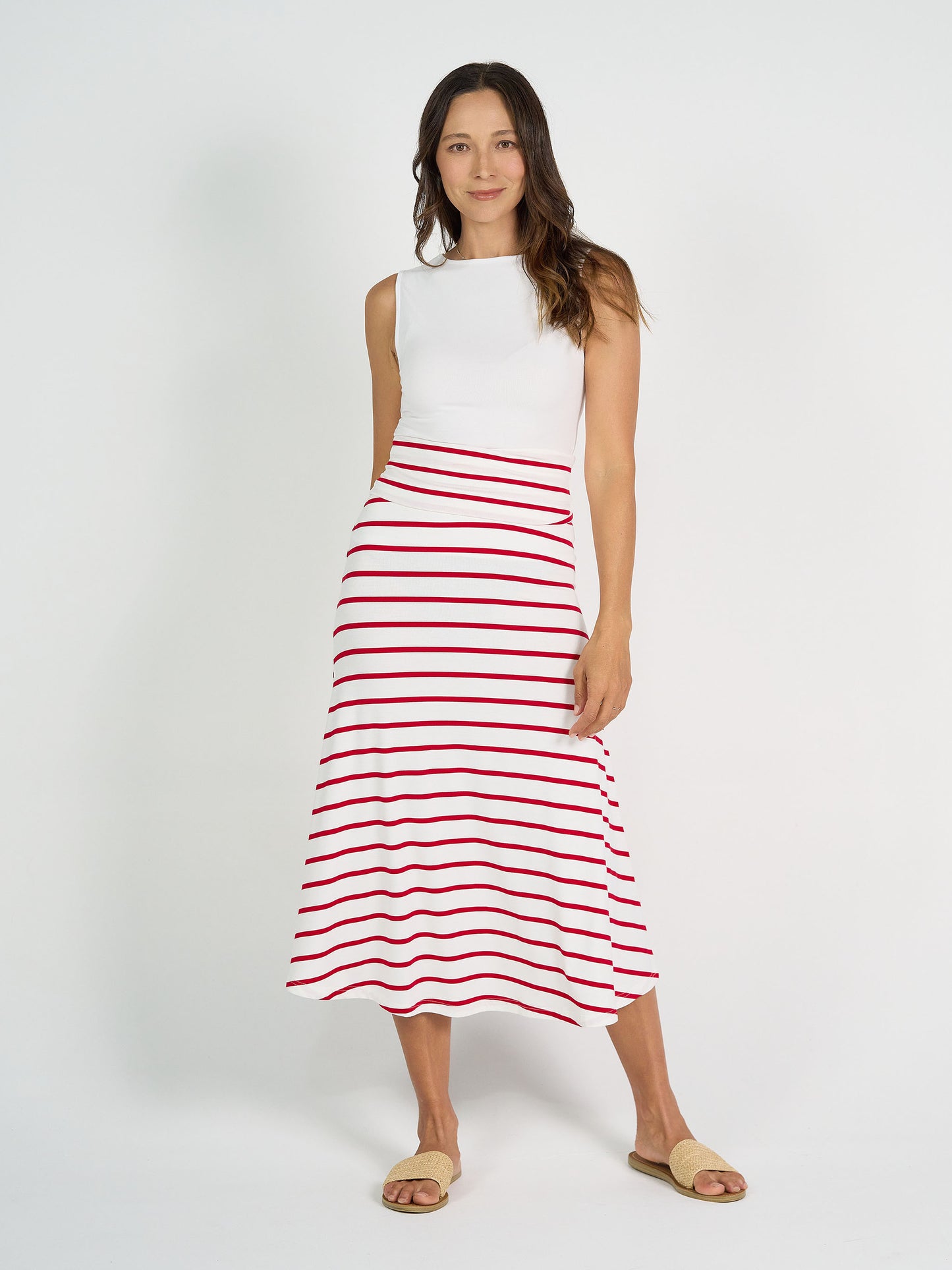 A woman stands against a white background wearing the Kate Bamboo Tank and a flowy white skirt with red stripes. She pairs the outfit with beige sandals, and her long brown hair is styled loose.