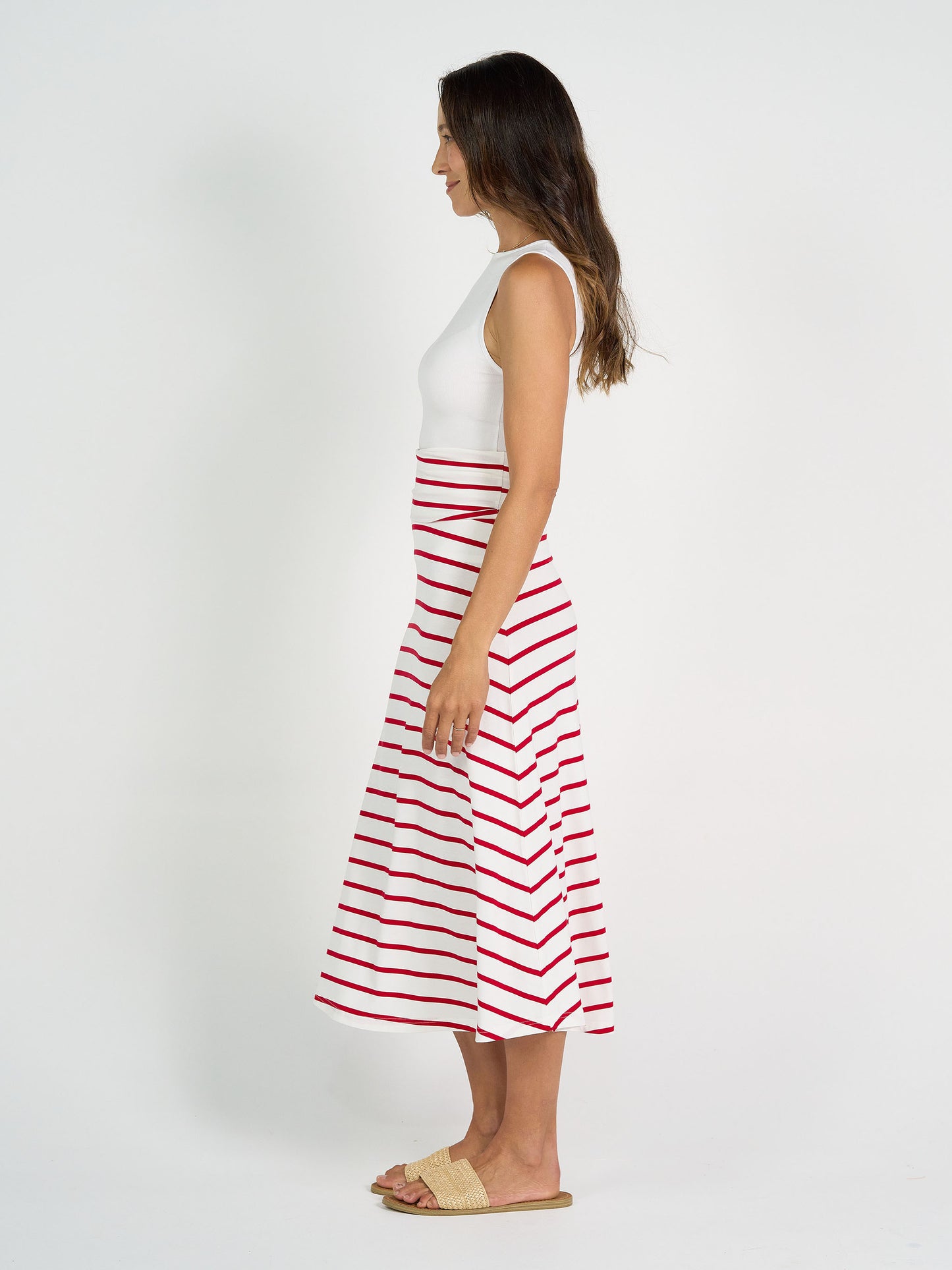 A woman stands in profile against a white background, smiling softly. She wears a sleeveless white top, the Jackson Bamboo Maxi Skirt in Lipstick & White Stripe, and beige woven sandals. Her long hair is down.