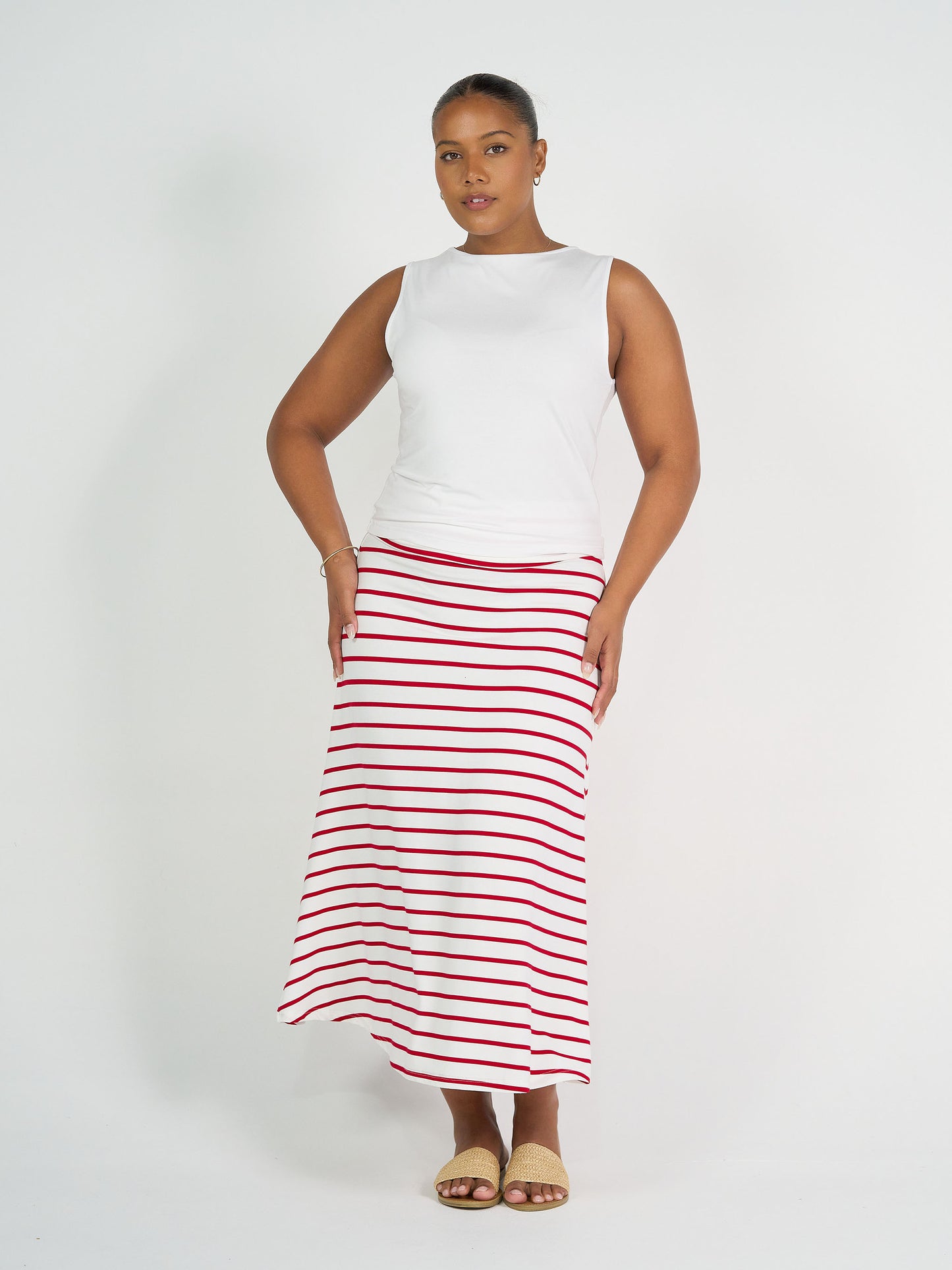 A woman poses against a white background wearing a sleeveless white top, tan sandals, simple jewelry, and the Jackson Bamboo Maxi Skirt in Lipstick & White Stripe.