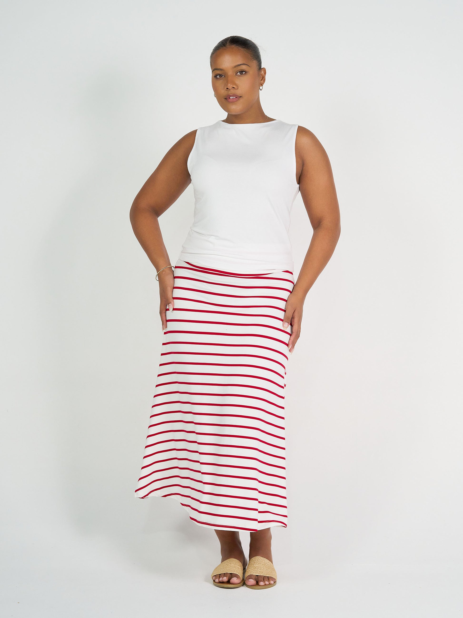 A woman poses against a white background wearing a sleeveless white top, tan sandals, simple jewelry, and the Jackson Bamboo Maxi Skirt in Lipstick & White Stripe.
