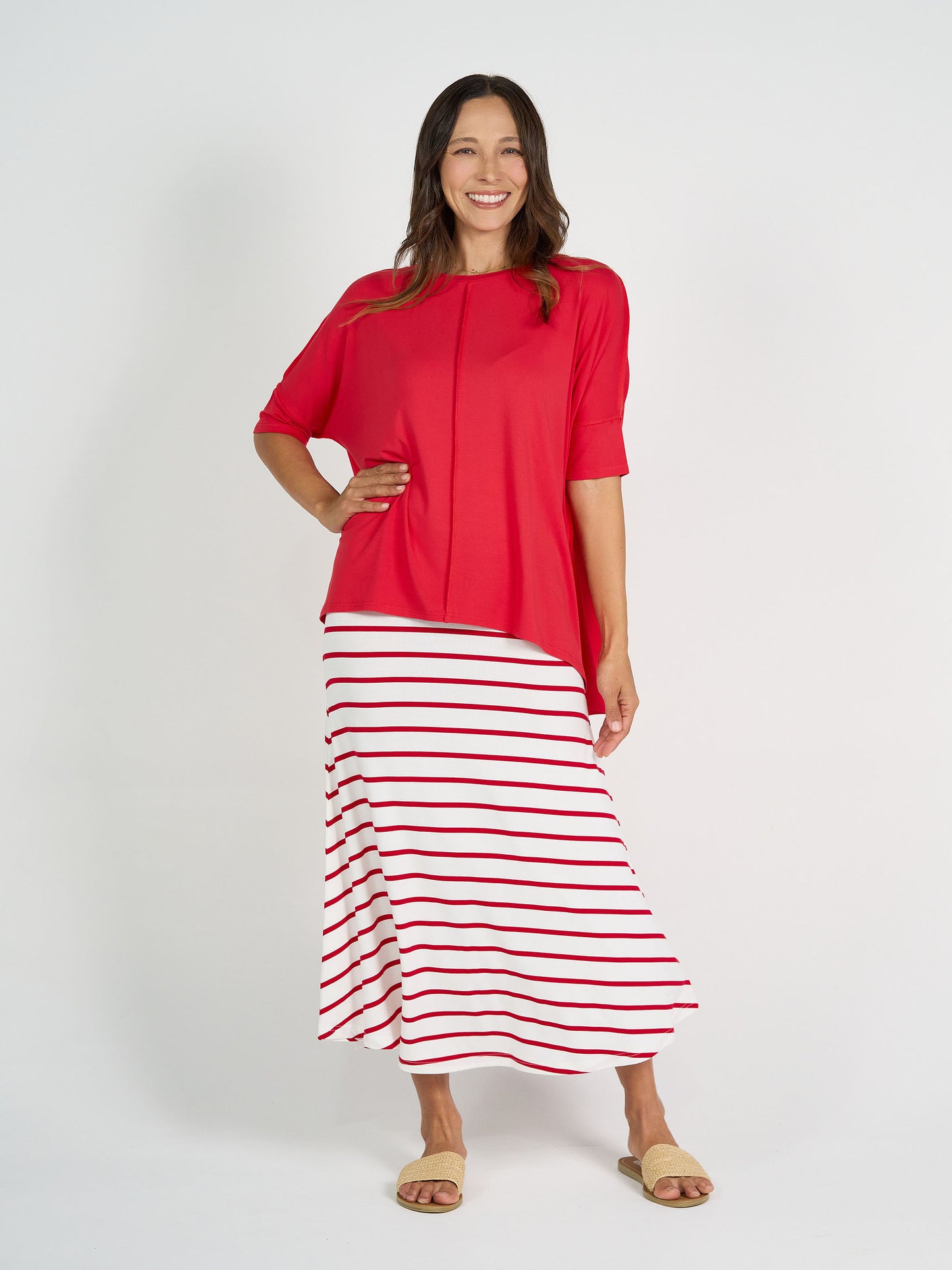 A woman smiles in a loose red top, beige slide sandals, and the Jackson Bamboo Maxi Skirt in Lipstick & White Stripe, standing against a plain white background.