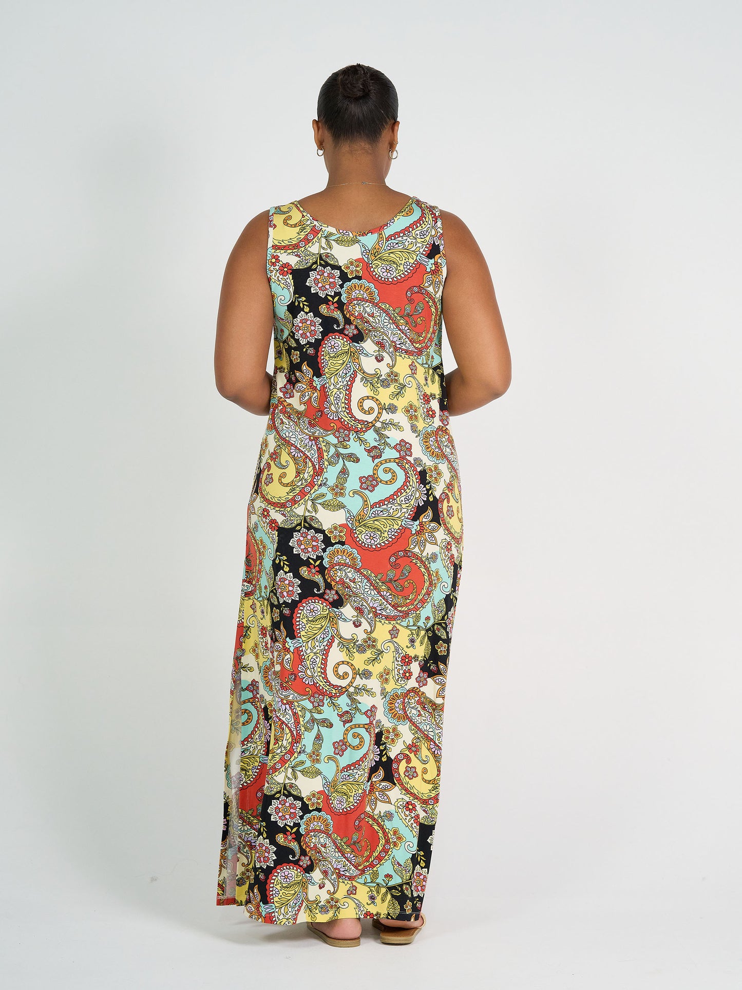 A person faces away, wearing the Janet Sleeveless Bamboo Maxi Dress - Noosa Pucci, featuring a colorful paisley pattern in red, yellow, black, and light blue. The background is plain white.