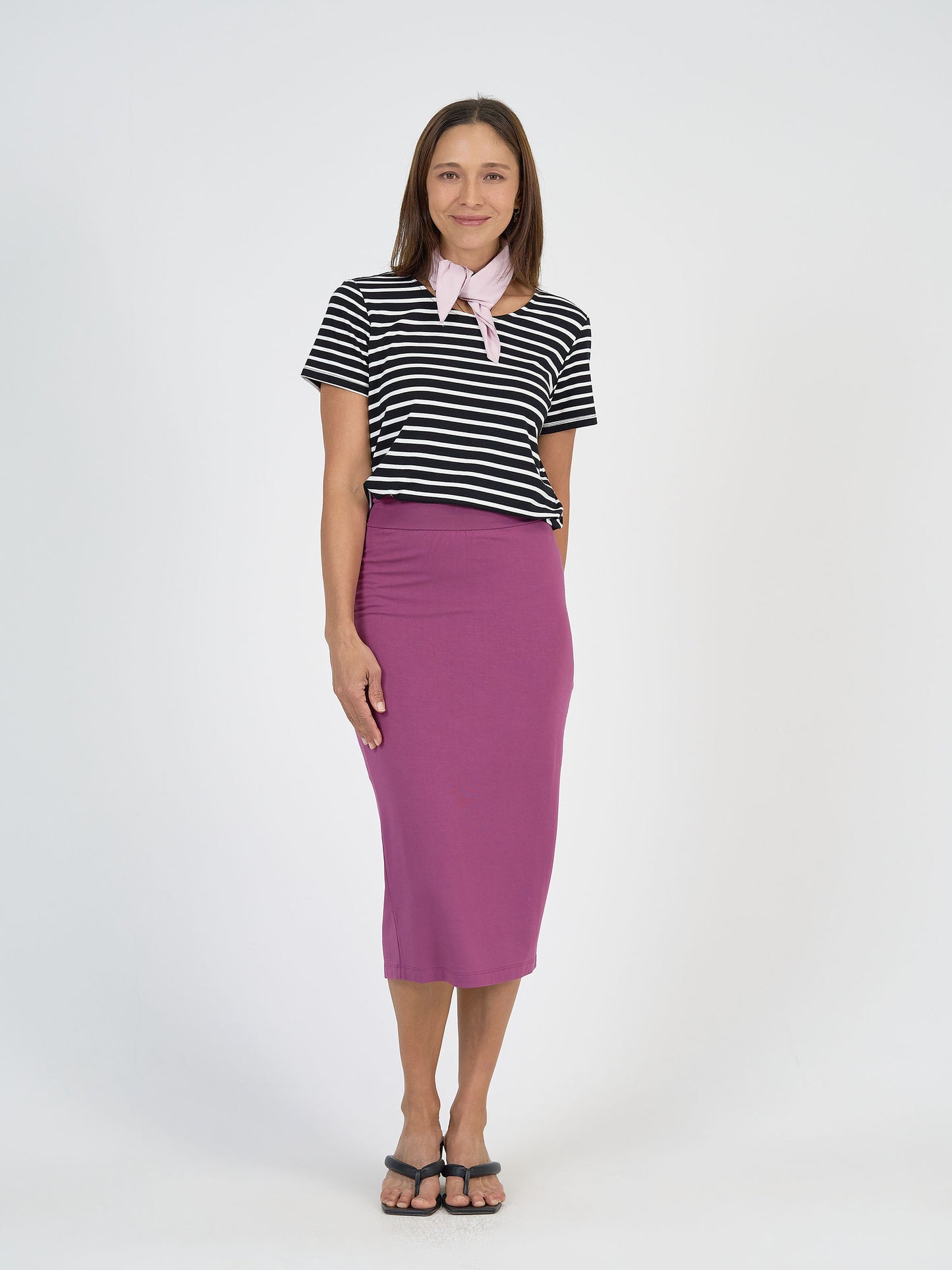 A woman with straight brown hair smiles against a white background, wearing a black and white striped t-shirt, purple midi skirt, black sandals, and the Benni Bamboo Scarf - Lilac—a sustainable accessory perfect for versatile styling.