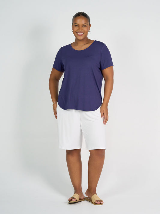 A woman stands smiling in front of a plain white background, wearing the Janis Short Sleeve Bamboo T-Shirt - Blue, white knee-length shorts, tan sandals, and hoop earrings.