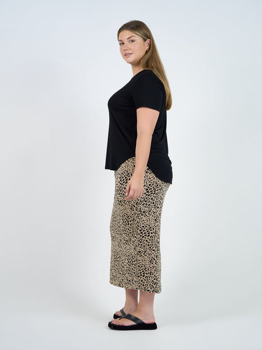 A woman stands sideways against a plain background, wearing the Janis Short Sleeve Bamboo T-Shirt in black, a beige and black animal print midi skirt, and black sandals. She has long straight hair and a neutral expression.
