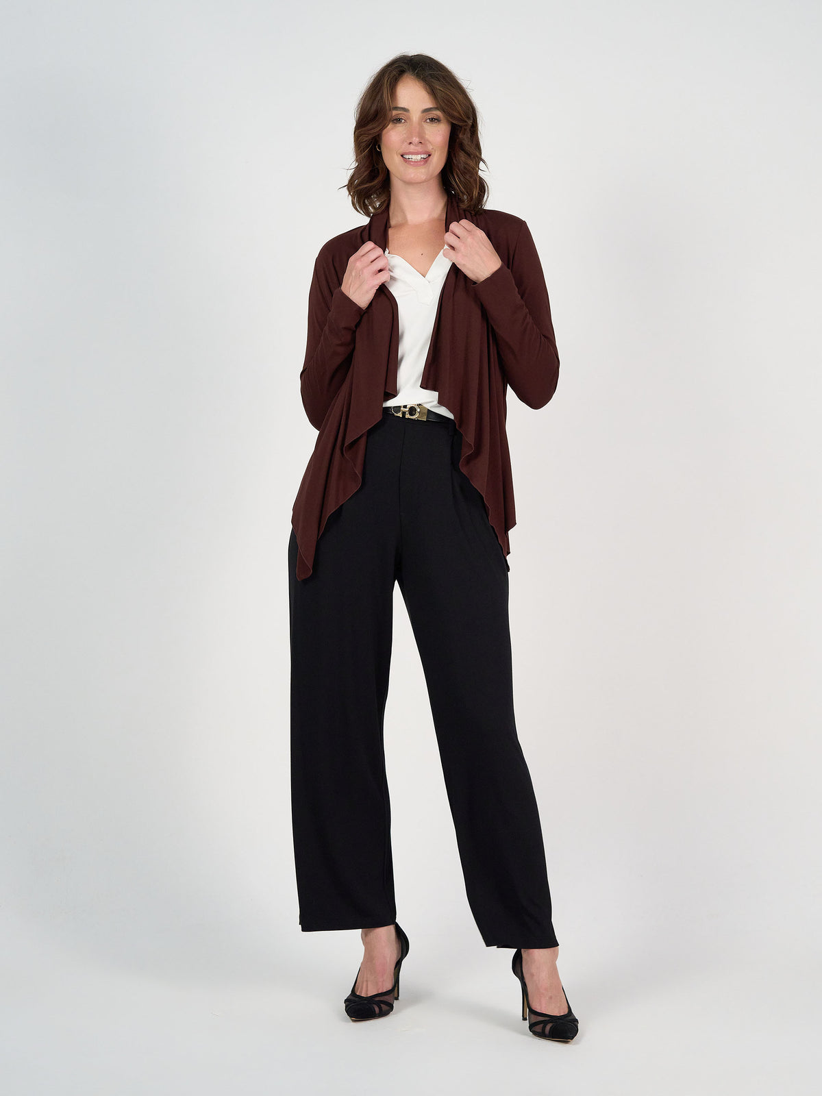 A woman stands and smiles against a plain background, wearing a Joni Bamboo Waterfall Cardigan/Jacket in Burgundy over a white blouse, with black wide-leg trousers and black heels. One hand touches her cardigan, the other rests at her side.