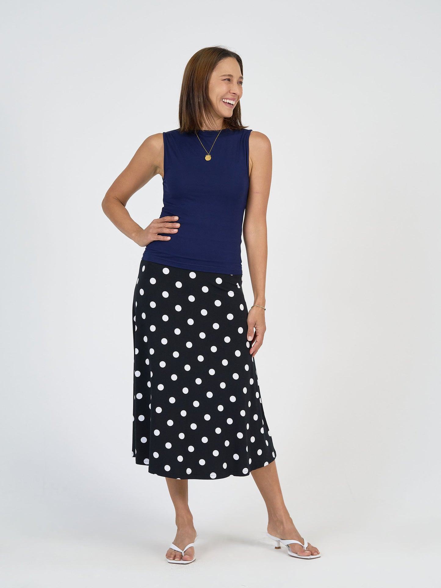 A woman stands smiling, wearing a sleeveless navy top, a black midi skirt with white polka dots, white sandals, and a gold necklace. She has straight brown hair and poses against a plain white background.