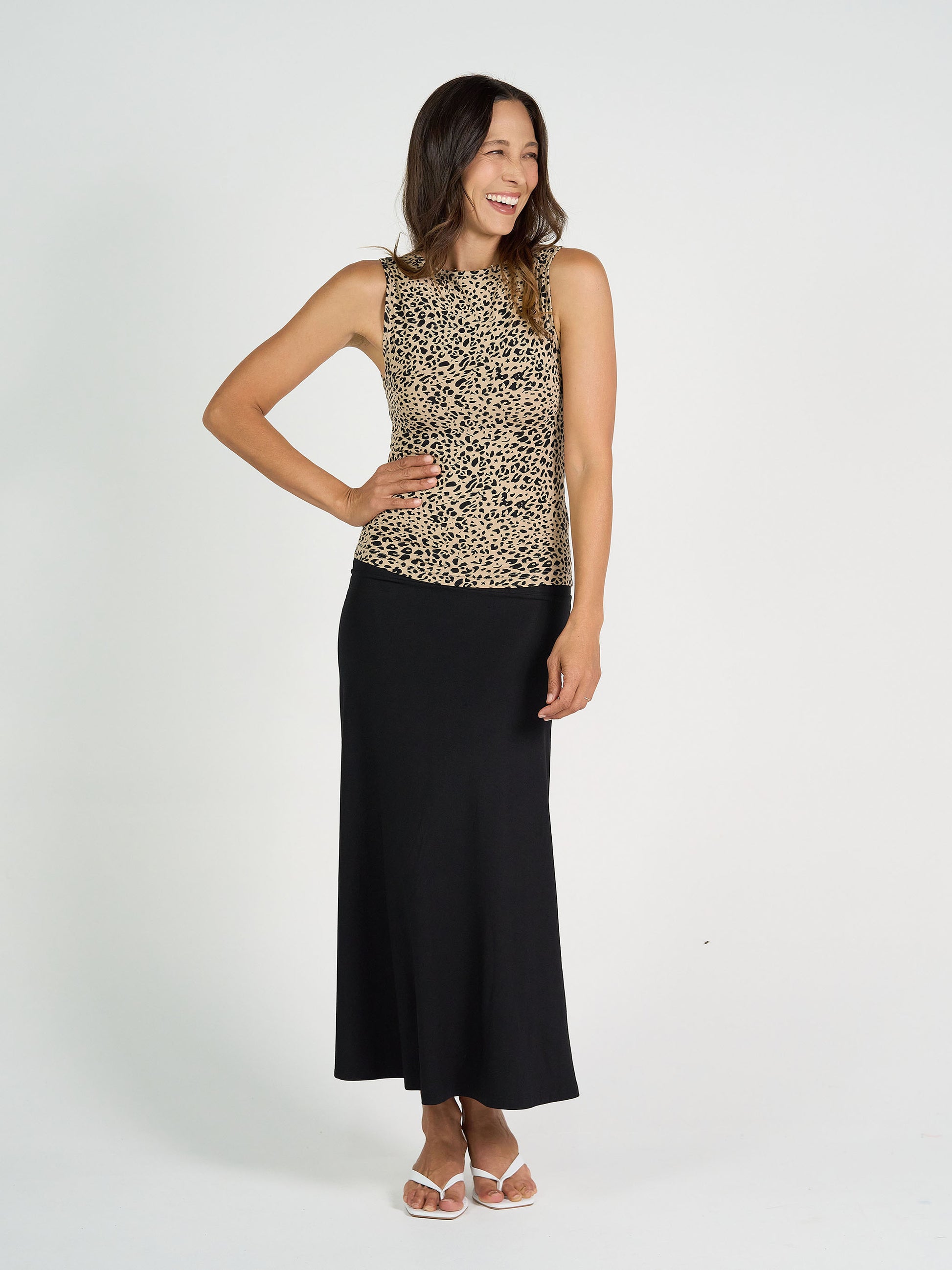 A woman smiles against a white background, wearing the Kate Bamboo Tank in leopard print with a long black skirt and white sandals. One hand rests on her hip, showcasing women's bamboo basics style.