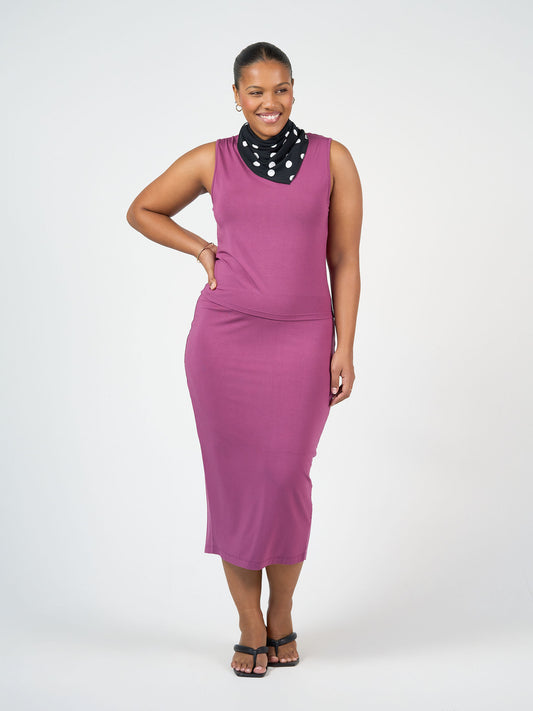 A woman poses confidently against a plain background, smiling in the Maxi Bamboo Whitney Tube Skirt - Plum, paired with black sandals and a black scarf with white polka dots, one hand on her hip and the other relaxed at her side.