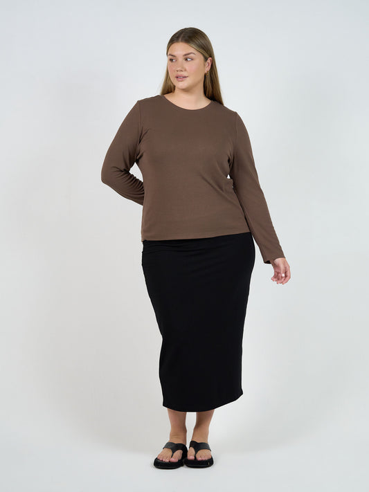 A woman stands against a white background wearing the Kelly Ribbed Long Sleeve Bamboo Top in Chocolate. She pairs it with a black midi skirt and sandals, her straight shoulder-length hair styled simply as she looks to the side in a relaxed pose.