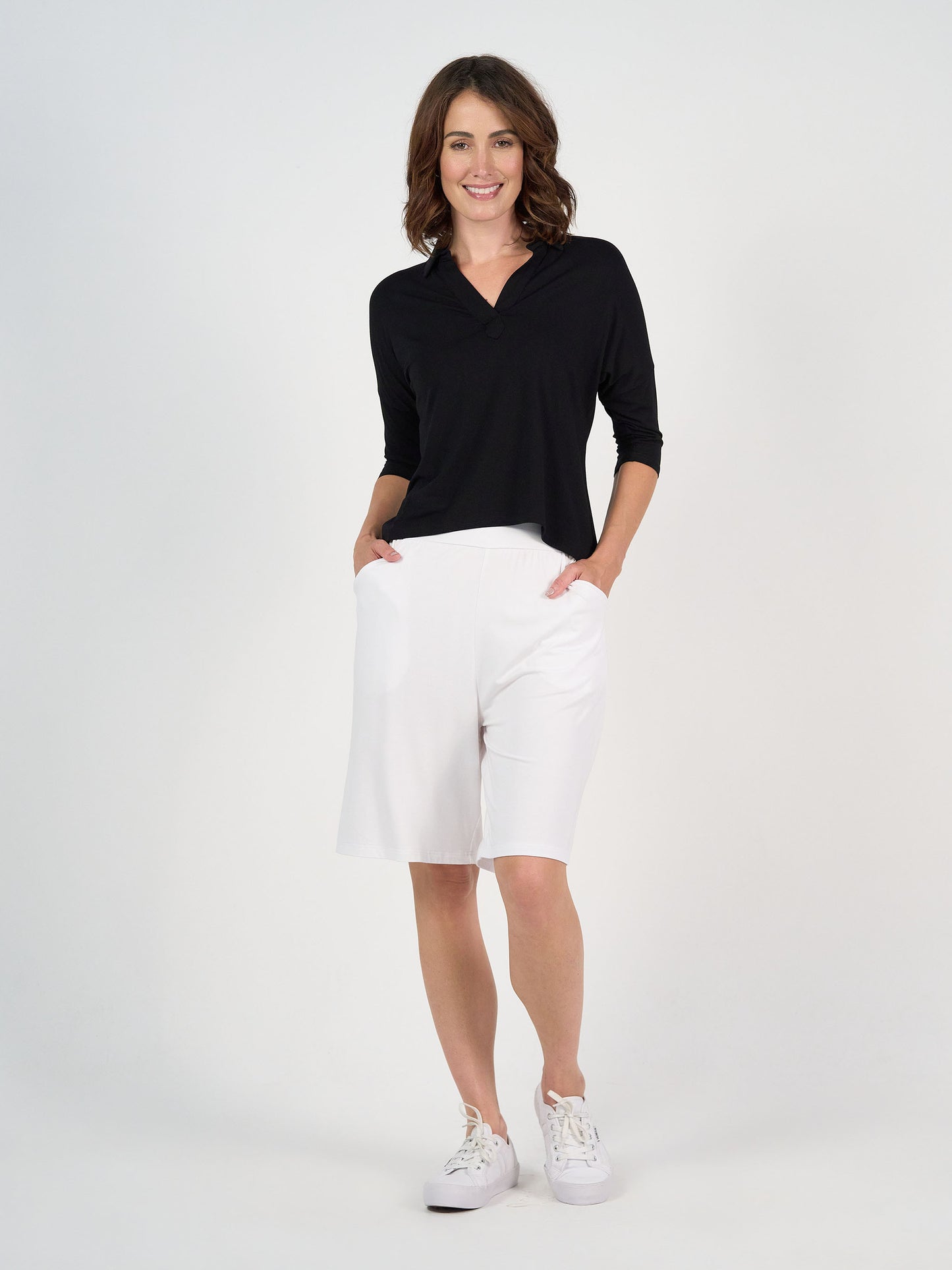 A woman stands against a plain white background, smiling. She is wearing the Khan Bamboo Collared Polo Top in black, white knee-length shorts, and white sneakers, with her hands in her pockets.