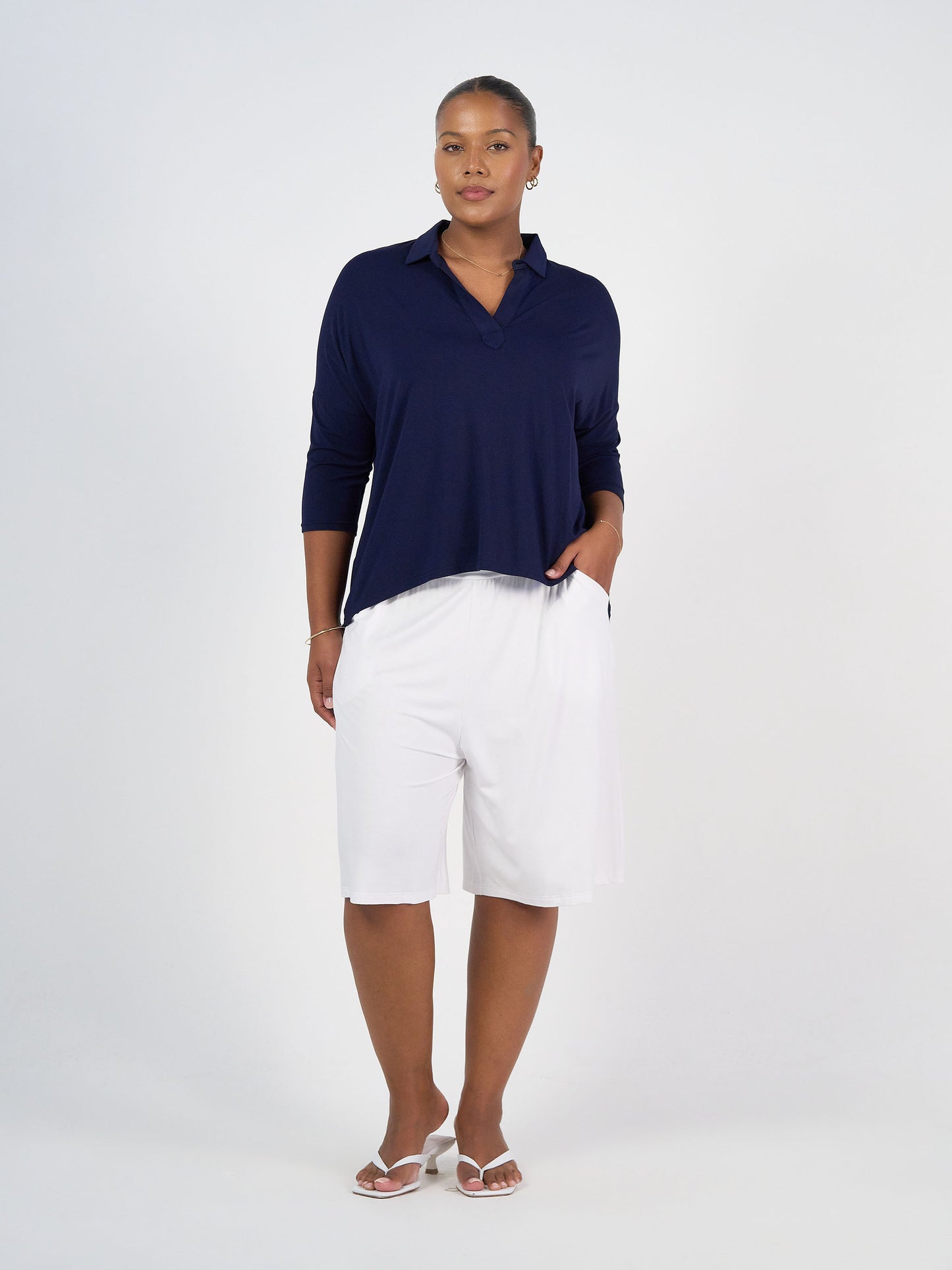 A woman stands against a plain white background, wearing the Khan Bamboo Collared Polo Top in navy, white knee-length shorts, and white sandals. Her right hand is in her pocket as she looks confidently at the camera.
