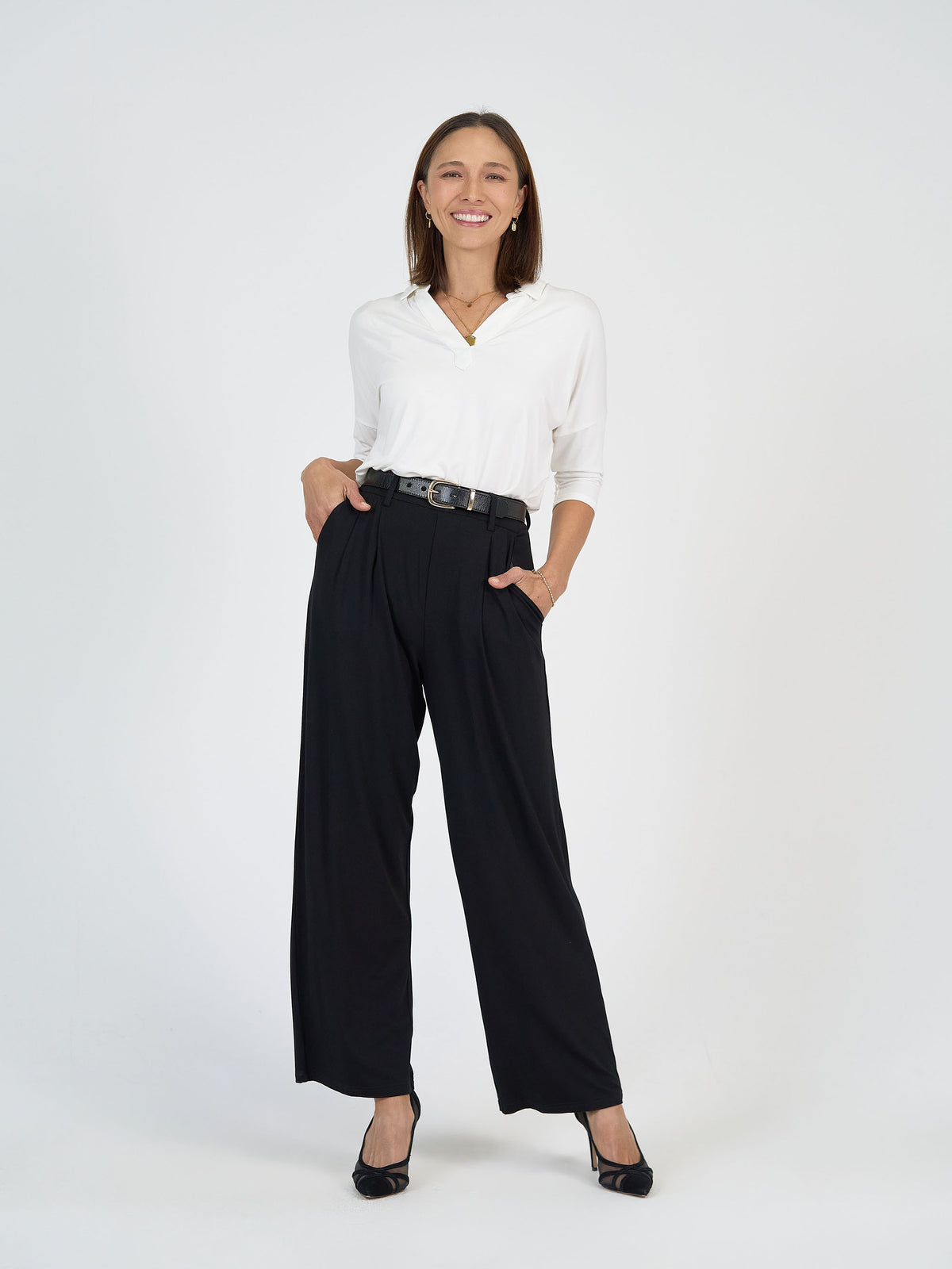 A woman stands against a plain white background, smiling with one hand in her pocket. She is wearing a white blouse, high-waisted black wide-leg pants, a black belt, and black heels.