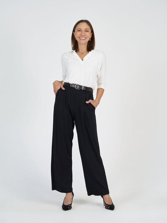 A woman stands against a plain white background, smiling with one hand in her pocket. She is wearing a white blouse, high-waisted black wide-leg pants, a black belt, and black heels.