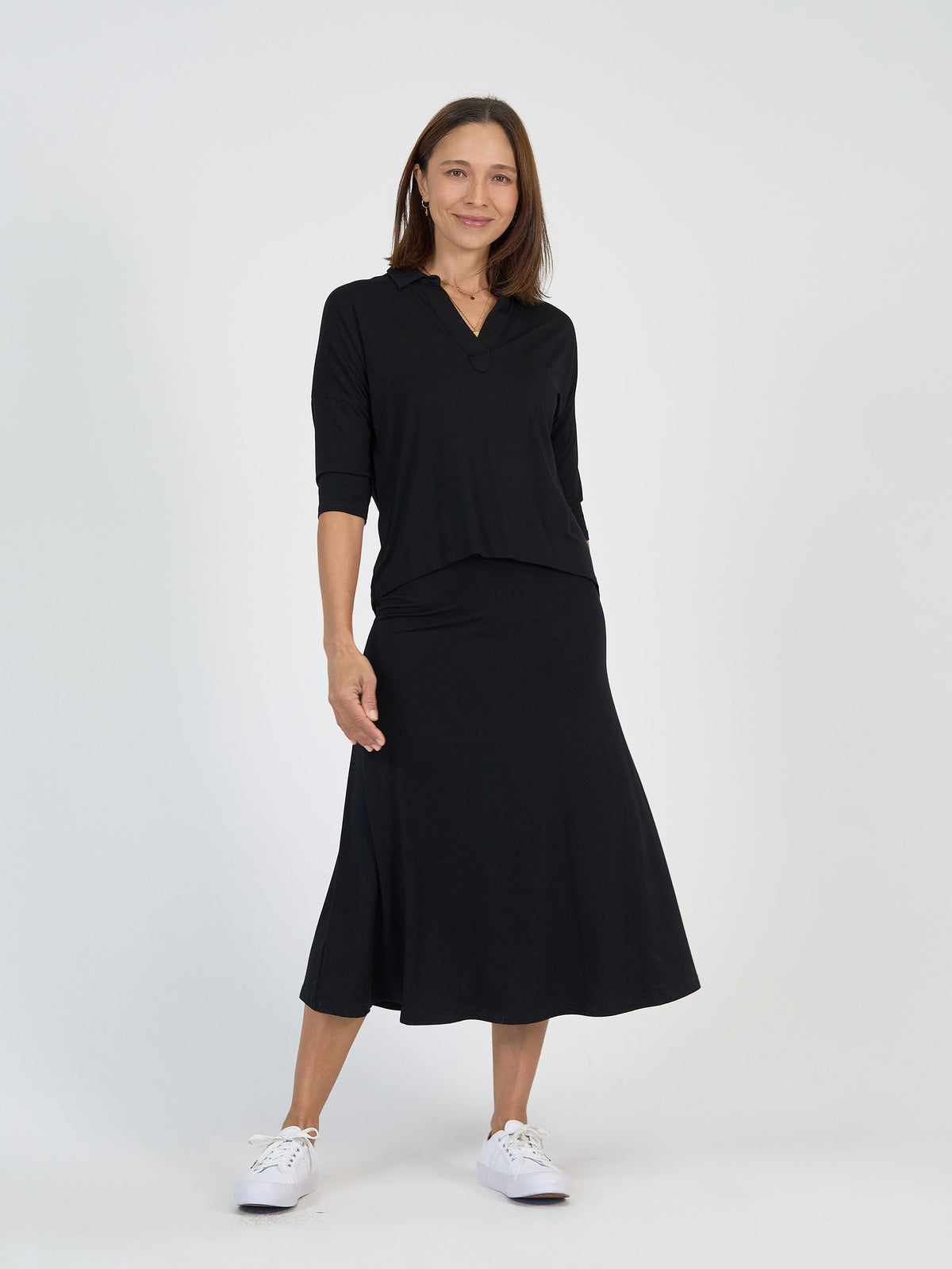 A woman stands against a plain white background, wearing a black long-sleeve top with a collar and a matching black midi skirt. She is smiling, with one hand by her side, and is wearing white sneakers.