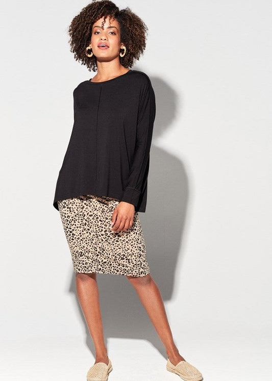 A woman poses against a light gray background, wearing the Stella Longsleeve Oversized Bamboo Slouch Top in black, paired with a knee-length leopard print skirt, woven beige slip-on shoes, and large hoop earrings. One leg is slightly bent.