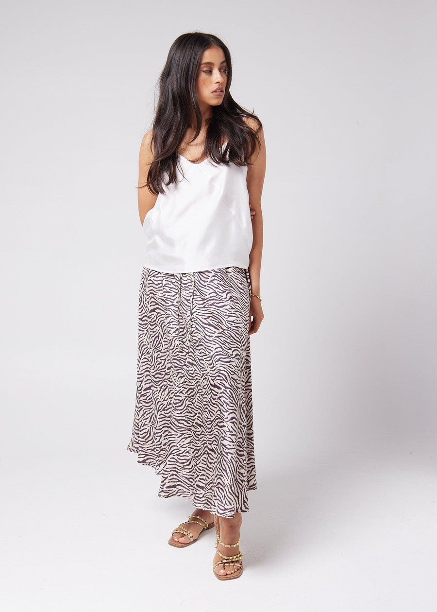 A woman looking sideways, showing off a mysterious vibe while wearing a white sleeveless bamboo top and animal printed midi skirt with a pair of brown strap sandals