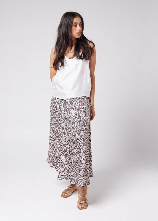 A woman looking sideways, showing off a mysterious vibe while wearing a white sleeveless bamboo top and animal printed midi skirt with a pair of brown strap sandals