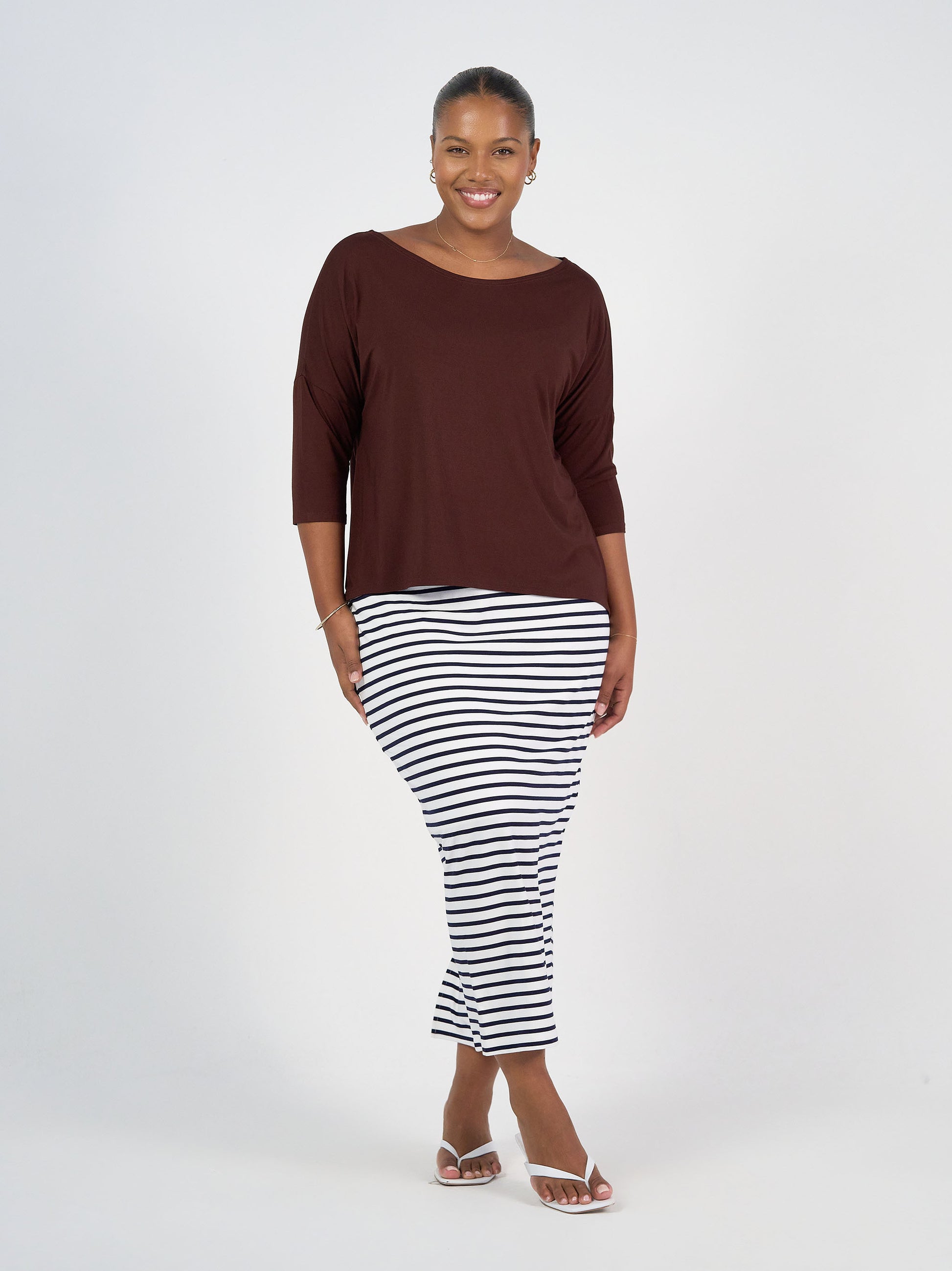 A woman smiles against a plain background, wearing a dark brown long-sleeve top, the Maxi Bamboo Whitney Tube Skirt with black and white stripes, and white sandals.