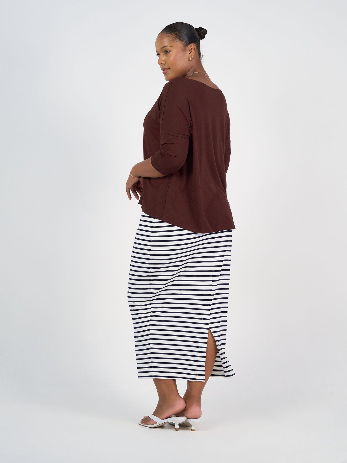 A woman stands sideways wearing a loose brown top with three-quarter sleeves and the Maxi Whitney Bamboo Tube Skirt in navy & white stripe, featuring a back slit. White heeled sandals complete her look, set against a plain white background.