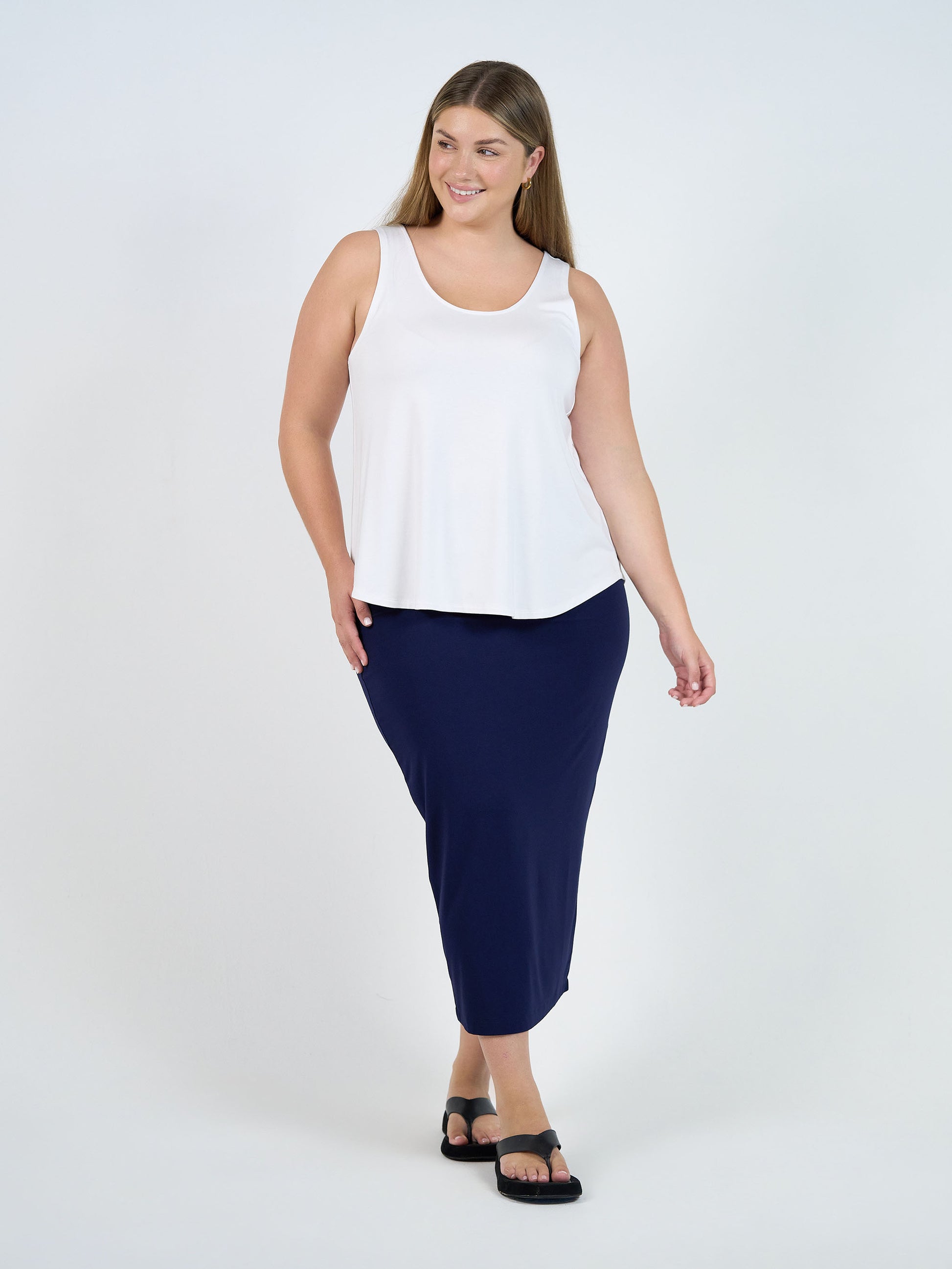 A woman with long hair wears a sleeveless white top, black sandals, and the Maxi Bamboo Whitney Tube Skirt in navy. She is smiling and walking against a plain white background.