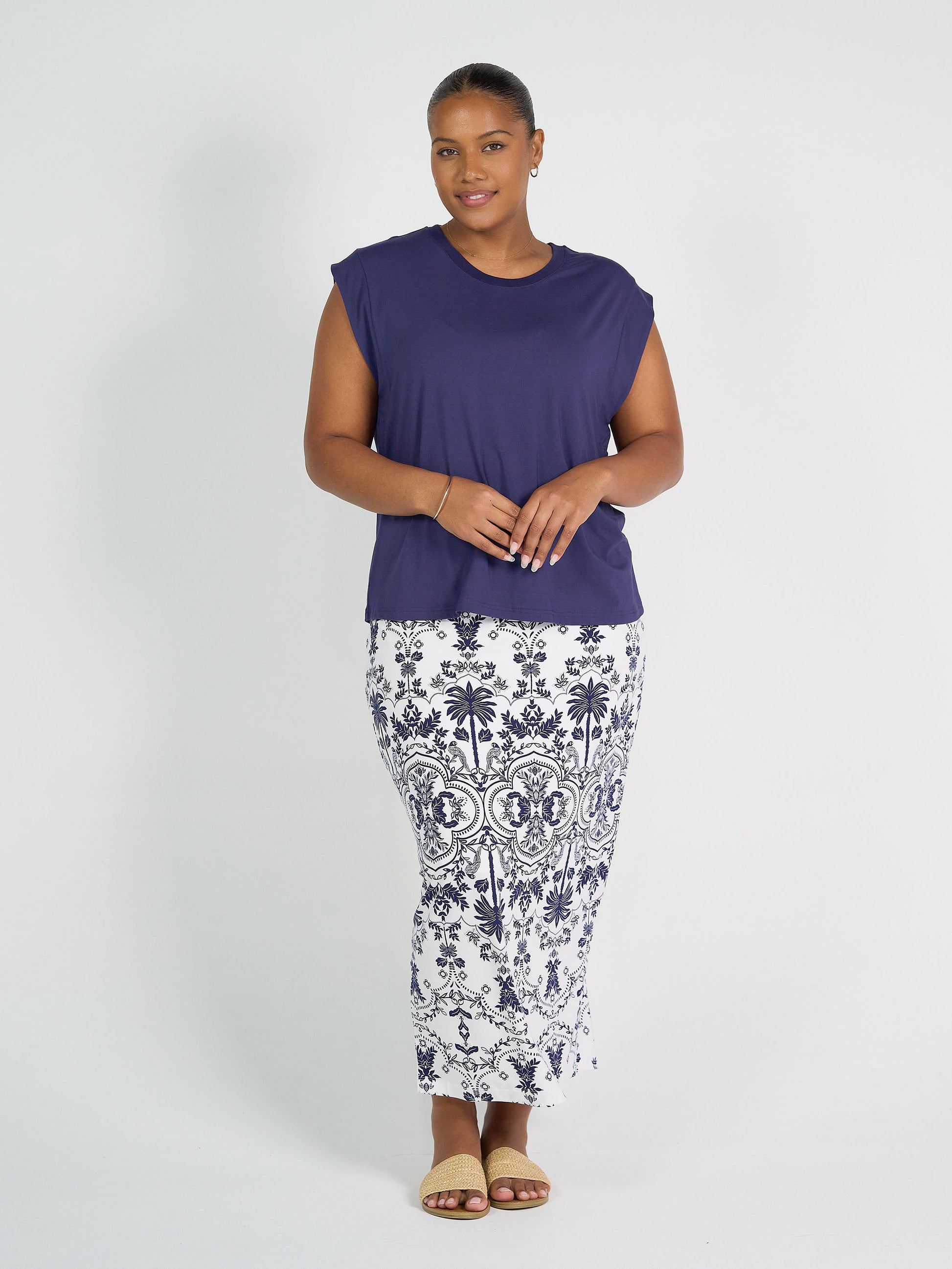 A woman stands against a white background, smiling, in a sleeveless navy top and the Maxi Bamboo Whitney Tube Skirt – Blue Palm, paired with tan sandals and simple jewelry. Her hands are loosely clasped.