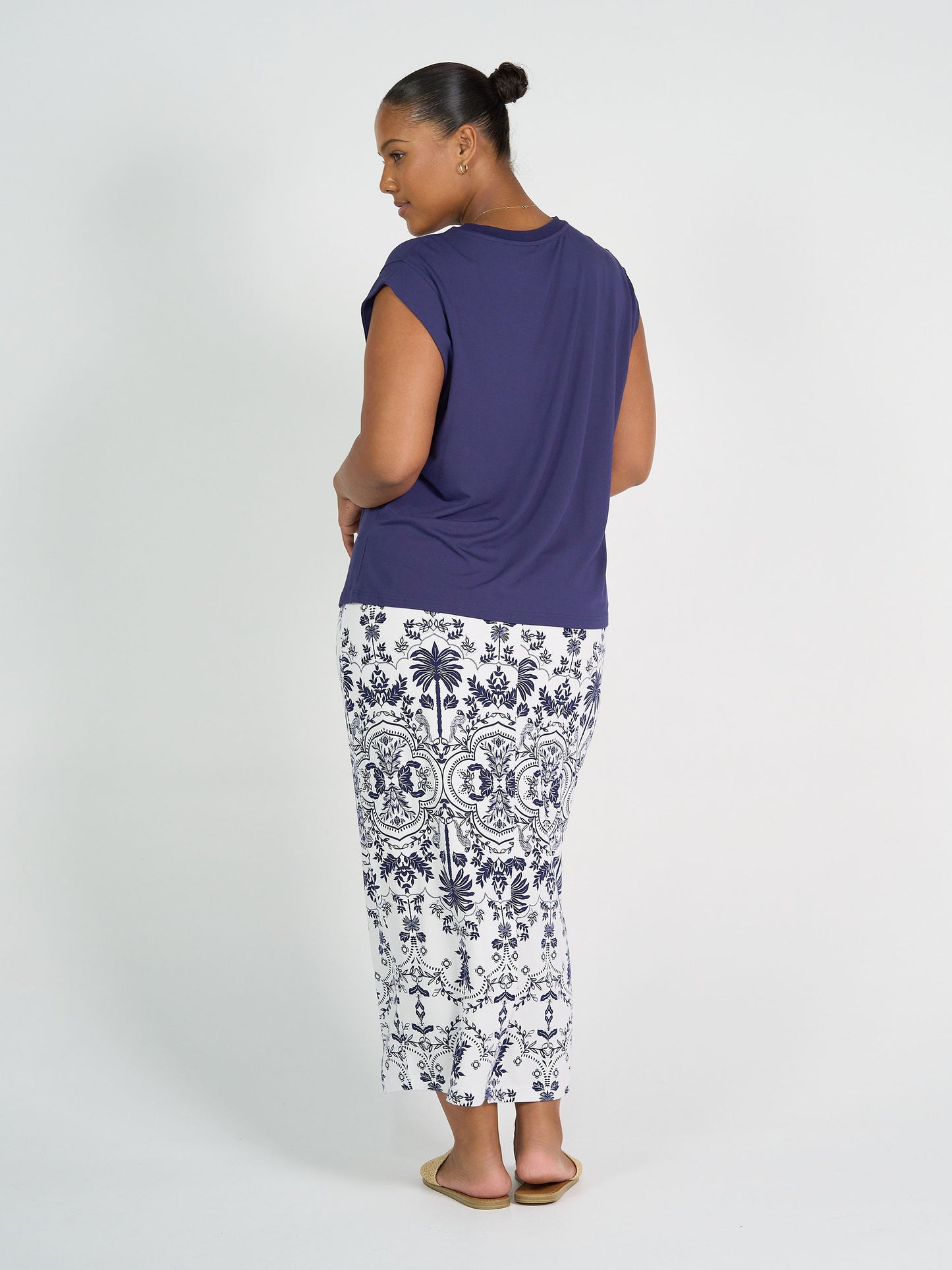 A woman with her hair in a bun faces away, wearing a navy short-sleeve top and the Maxi Bamboo Whitney Tube Skirt - Blue Palm, paired with flat sandals against a plain white background.