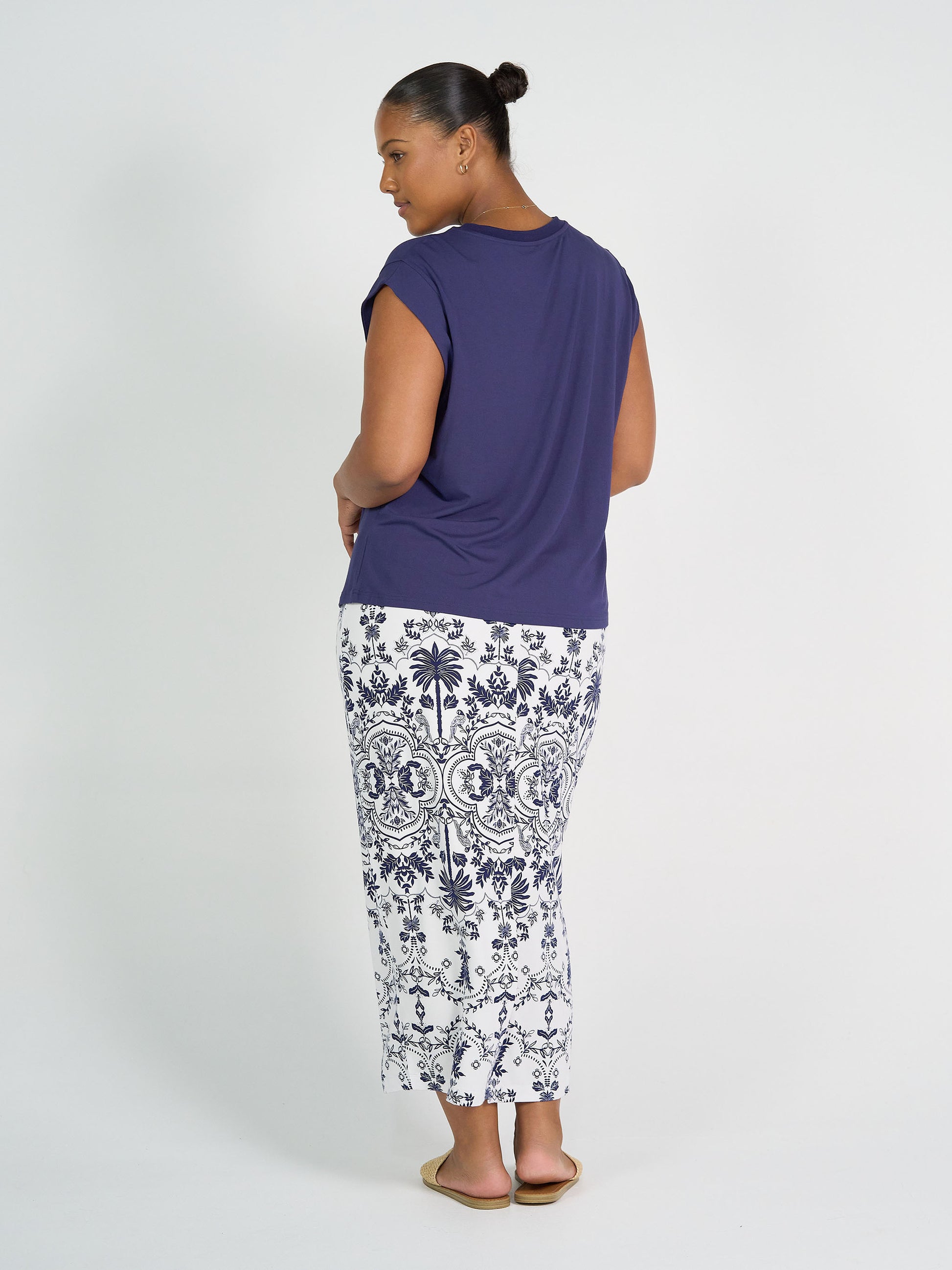 A woman with her hair in a bun faces away, wearing a navy short-sleeve top and the Maxi Bamboo Whitney Tube Skirt - Blue Palm, paired with flat sandals against a plain white background.