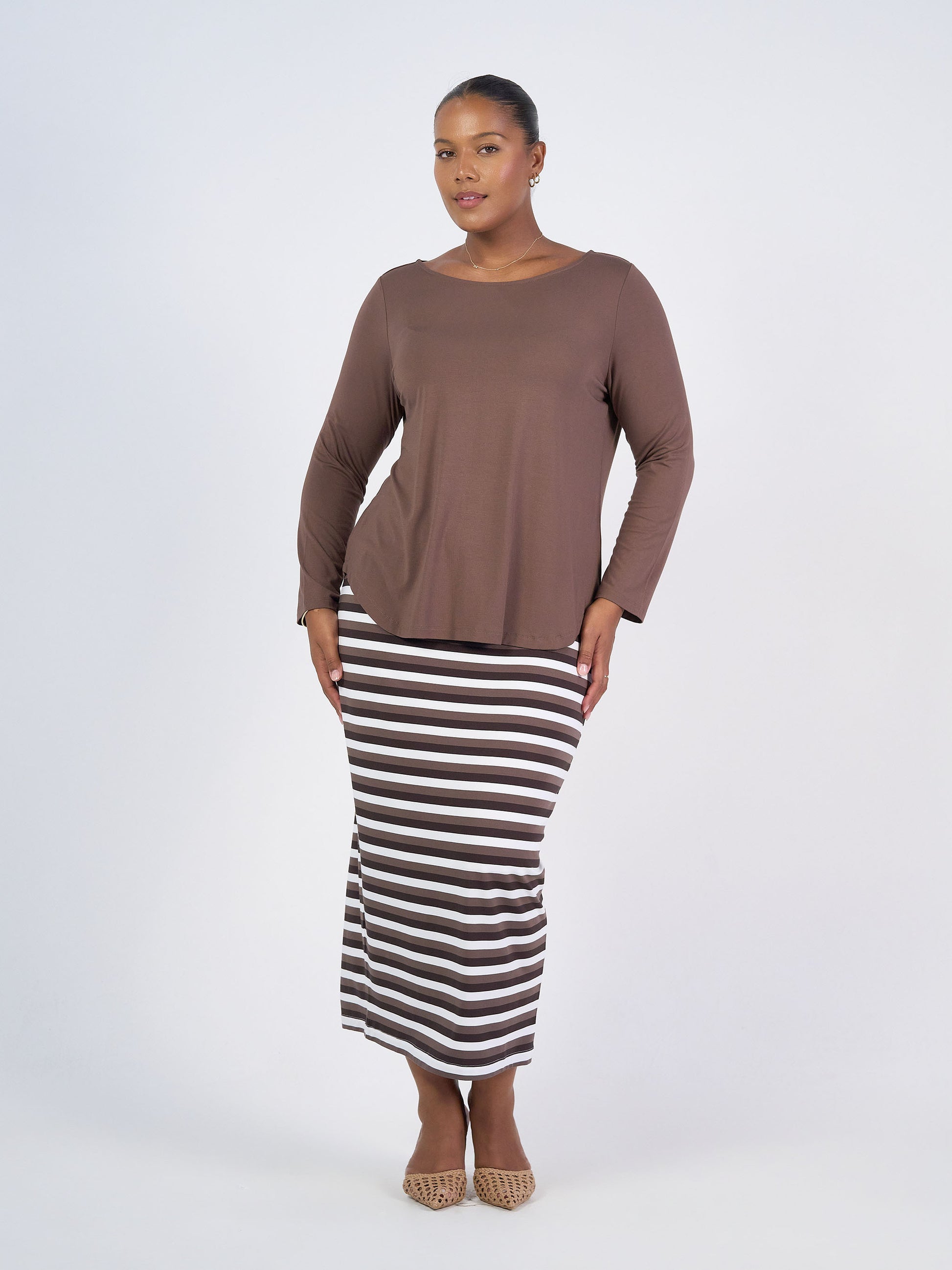 A woman stands against a white background, wearing the Adele Long Sleeve Bamboo Top with a black, white, and brown striped maxi skirt and beige pointed flats. She smiles softly and poses with her hands touching her skirt.