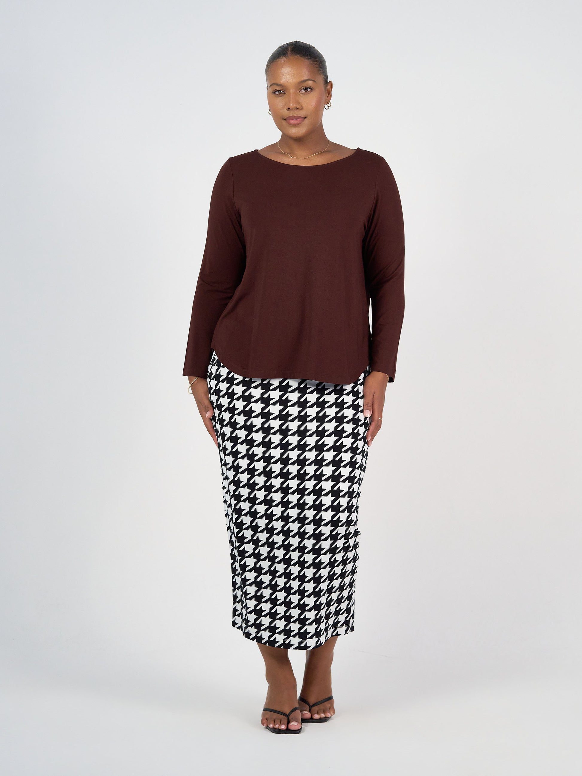 A woman with short hair stands against a plain background wearing the Adele Long Sleeve Bamboo Top in dark brown, paired with a high-waisted, black-and-white houndstooth ankle-length skirt and sandals.